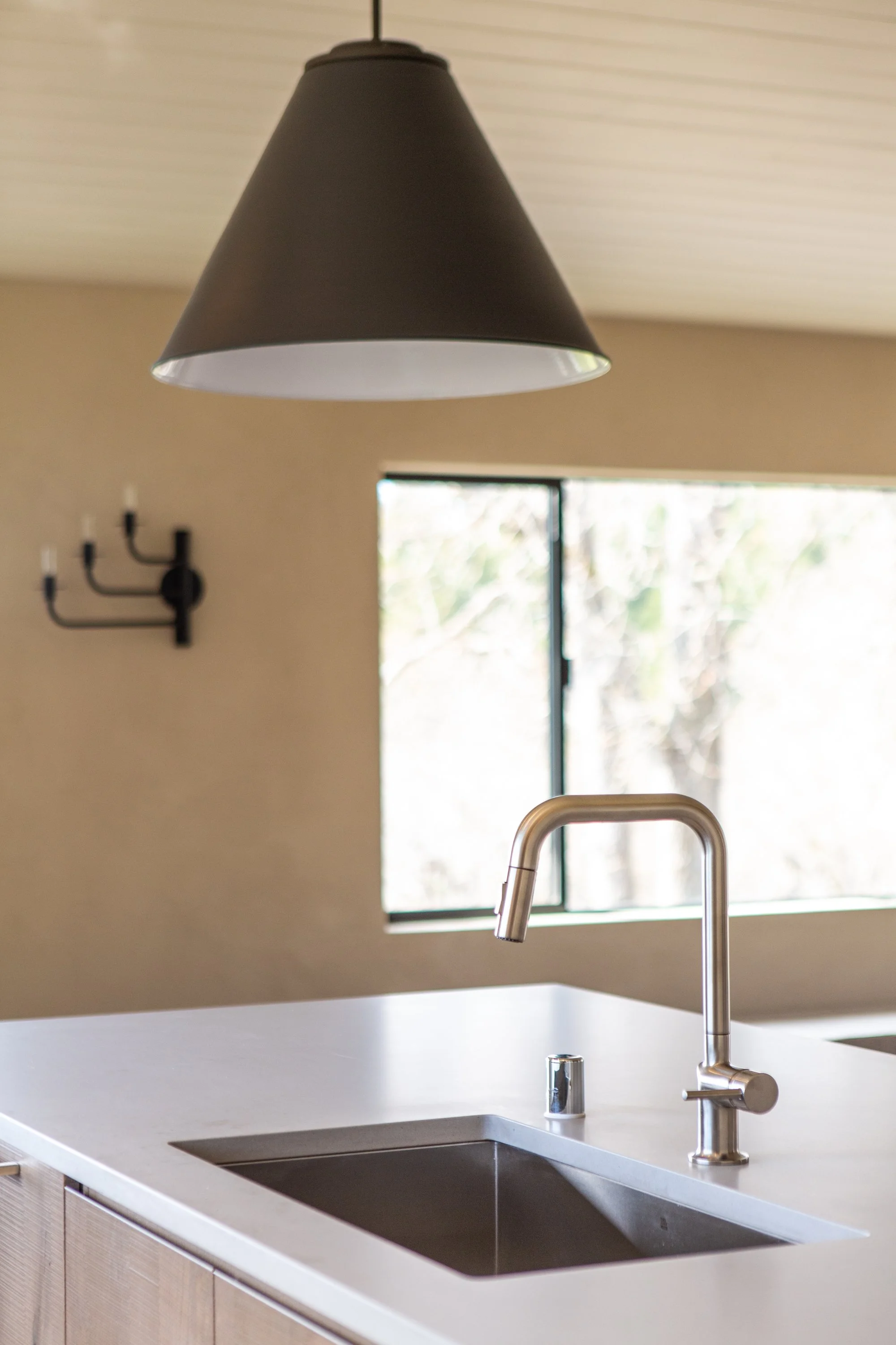 Modern kitchen sink with stainless steel faucet and beige cabinetry, window with blurred outdoor view, black wall-mounted candle holder, black pendant light hanging from ceiling.