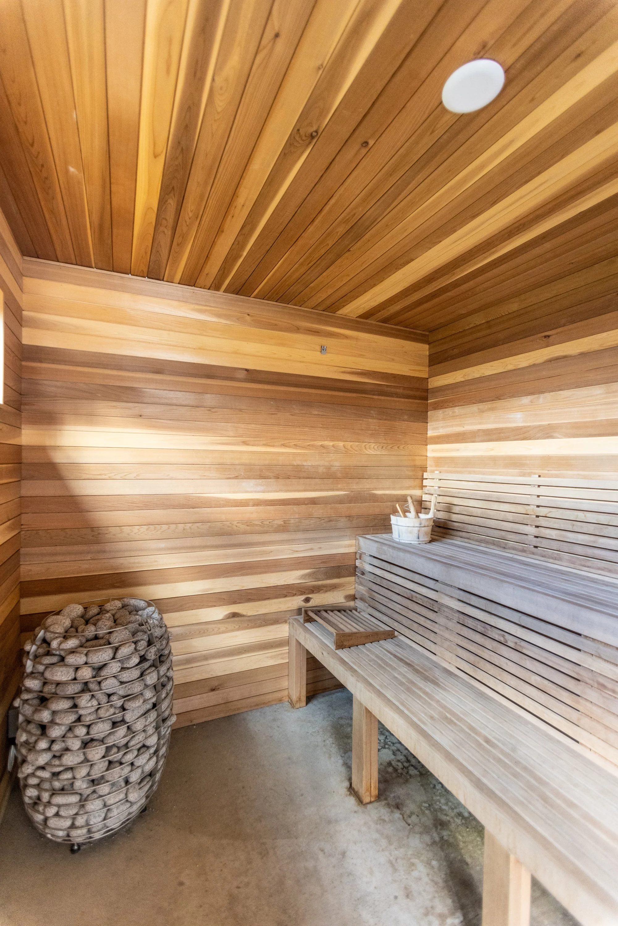 Wooden sauna interior with wooden bench, bucket, ladle, and a basket of stones.