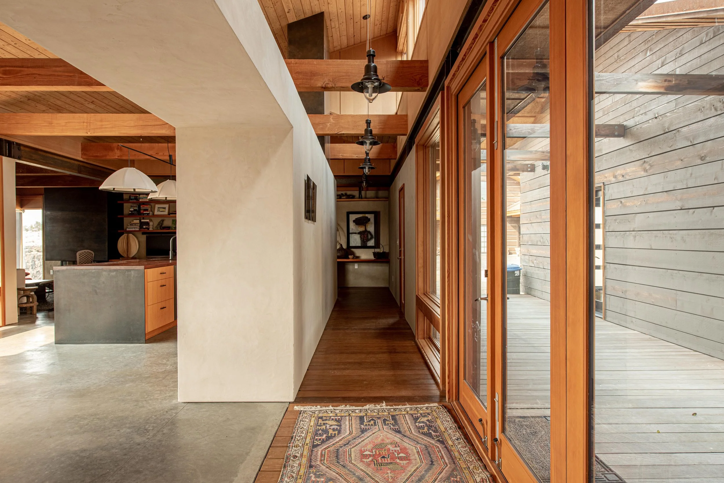 Interior view of a modern home showcasing a hallway with wooden framed glass doors leading outside, a colorful patterned rug on a wooden floor, and pendant and industrial-style ceiling lights.