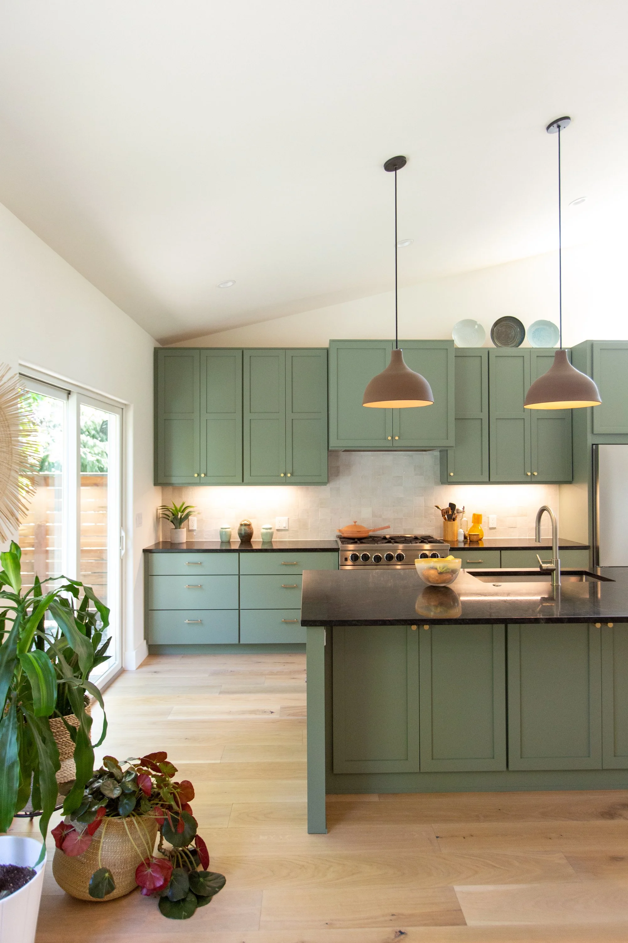 Modern kitchen with green cabinets, black countertop, beige tile backsplash, pendant lights, potted plants, and a bowl on the island.