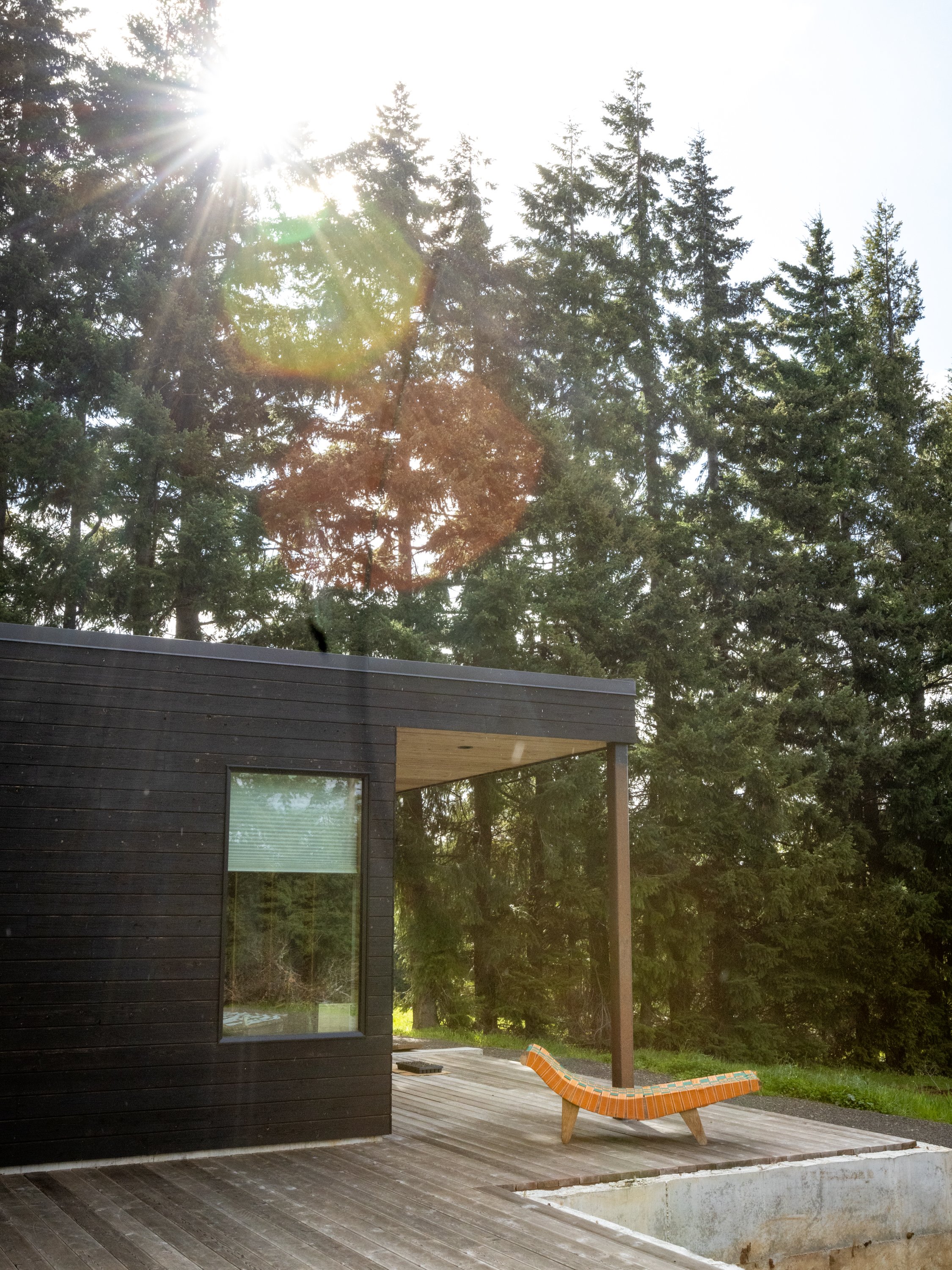 The image shows an outdoor patio area with a hot tub, a modern black wooden house with large windows, a sun lounger with a striped orange cushion, and tall pine trees in the background. The sun is shining brightly, creating lens flare and a warm glow