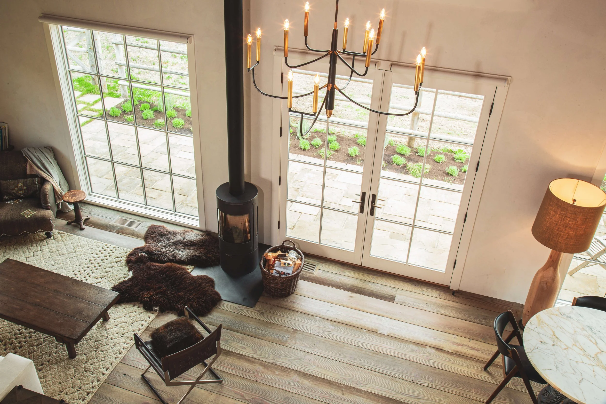 Living room with large glass doors opening to a garden, a wood stove, a chandelier, a sofa with throw blanket, a small side table, a basket with magazines, and a rug with fur animal hides.