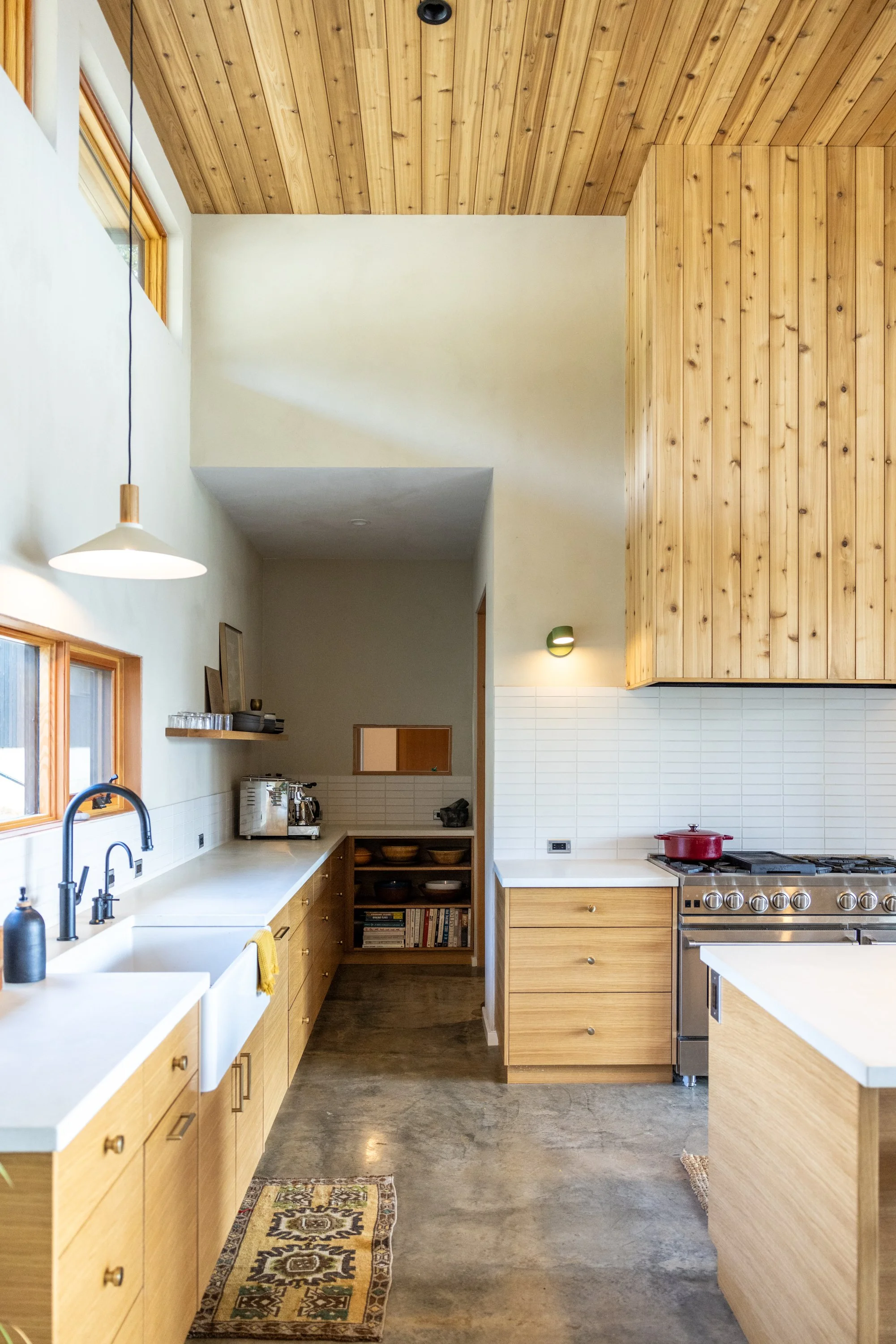 A modern kitchen with wooden cabinetry, a white countertop, a sink with a black faucet, and kitchen appliances including a stainless steel stove with a red pot. The ceiling and upper wall are covered with wood paneling, and there are windows allowing