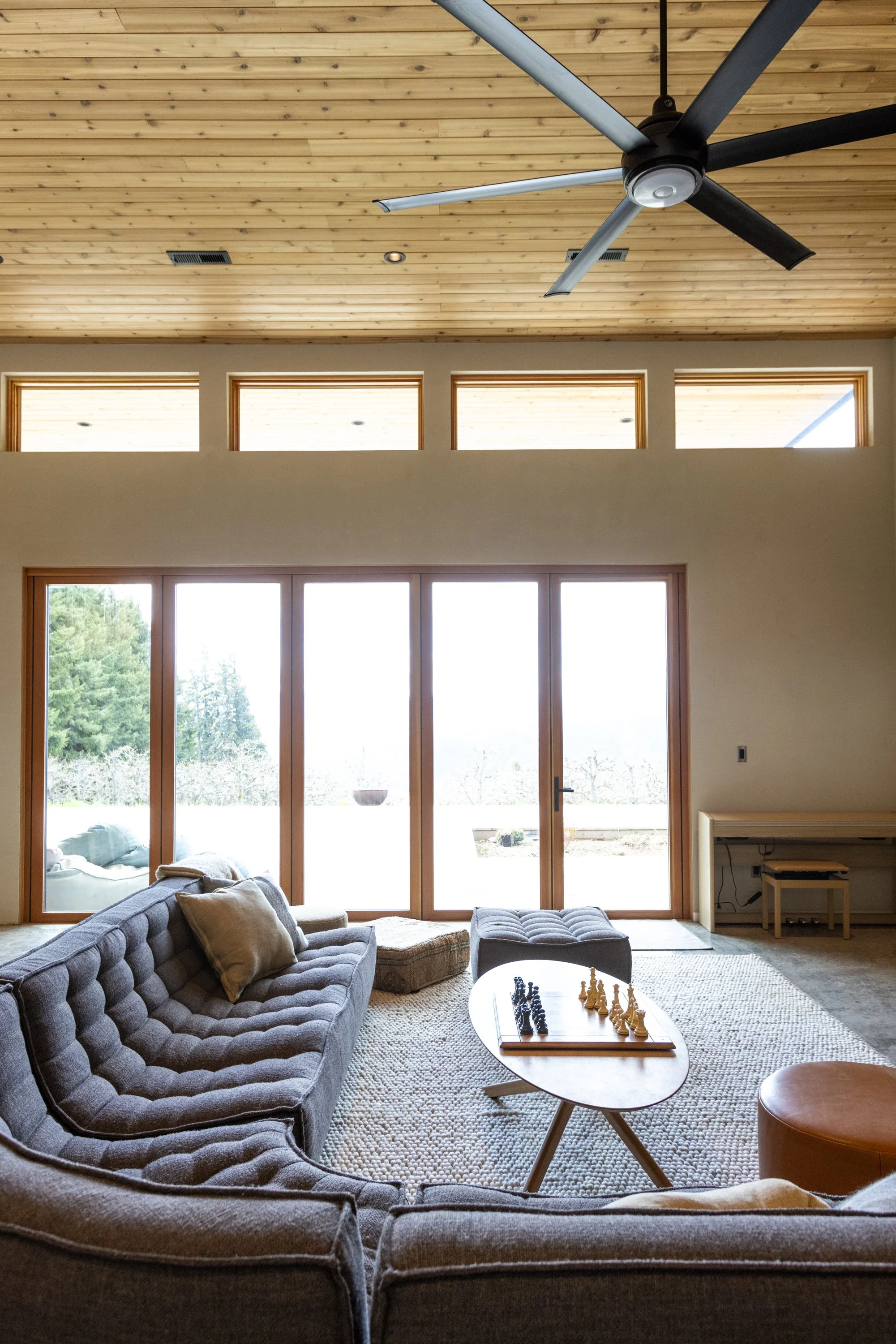 Living room with large glass doors, a gray tufted sofa with beige pillows, a round coffee table with a chess set, and a beige ottoman. Wooden ceiling and high windows letting in natural light, with trees visible outside.