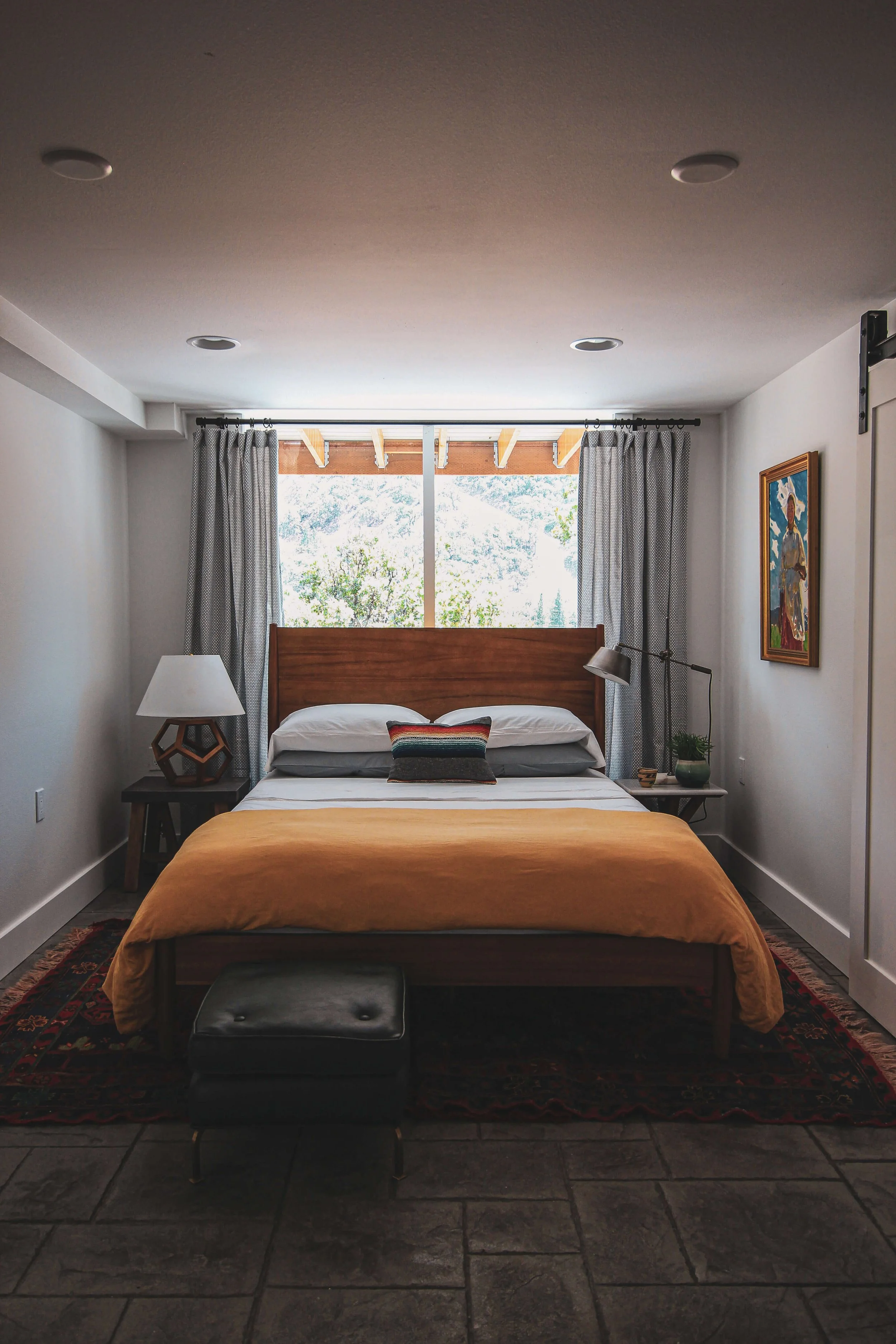 A bedroom with a bed covered in a mustard yellow blanket, a decorative pillow, and a wooden headboard. There are two bedside tables with lamps, one on each side of the bed. A large window with gray patterned curtains lets in natural light. There is a