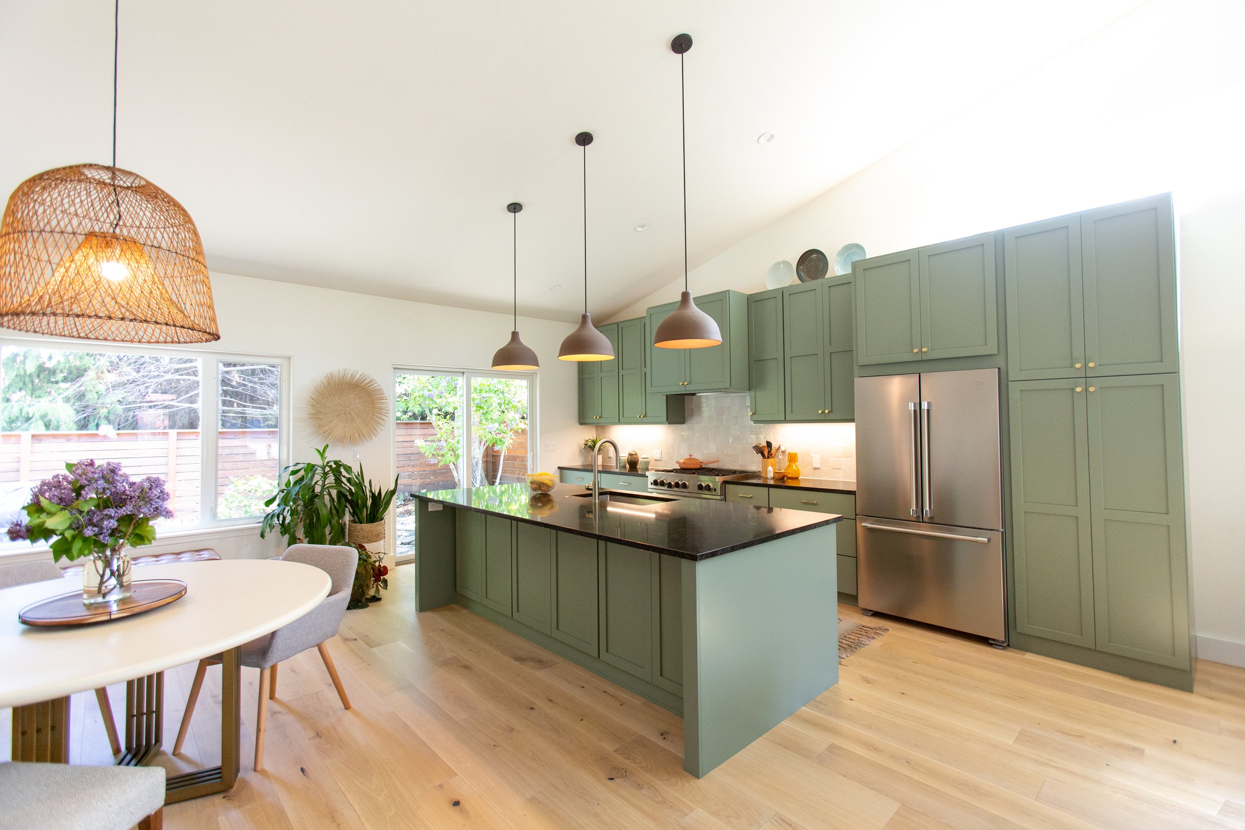 A modern kitchen with green cabinets, black countertops, and stainless steel appliances. Hanging pendant lights and a basket light fixture are visible, with a window showing trees outside and a dining area with a white table and chairs.