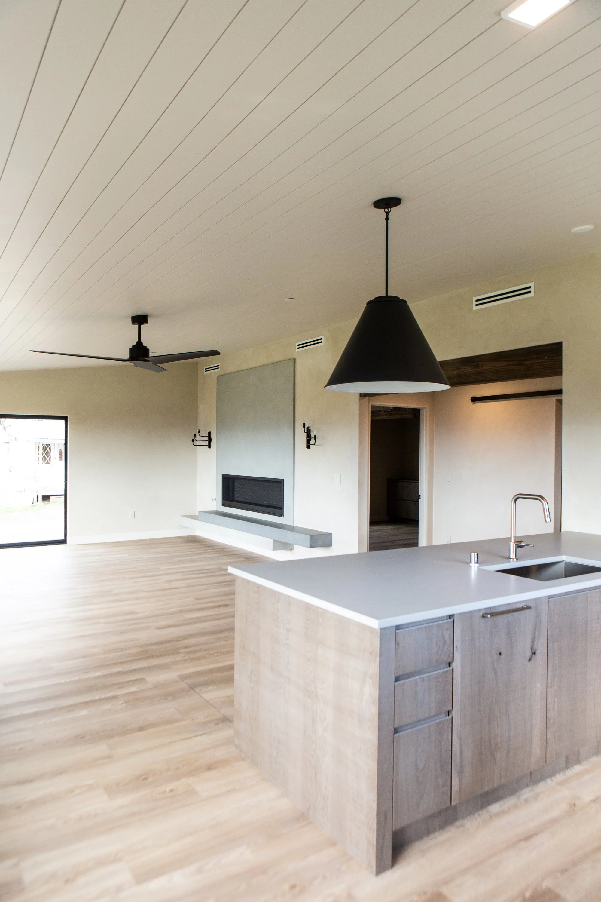Interior view of a modern open-concept kitchen with a white ceiling, light wood floors, a black ceiling fan, and a large black pendant light over a light wood kitchen island with white countertop. There is a fireplace on the wall and sliding glass do
