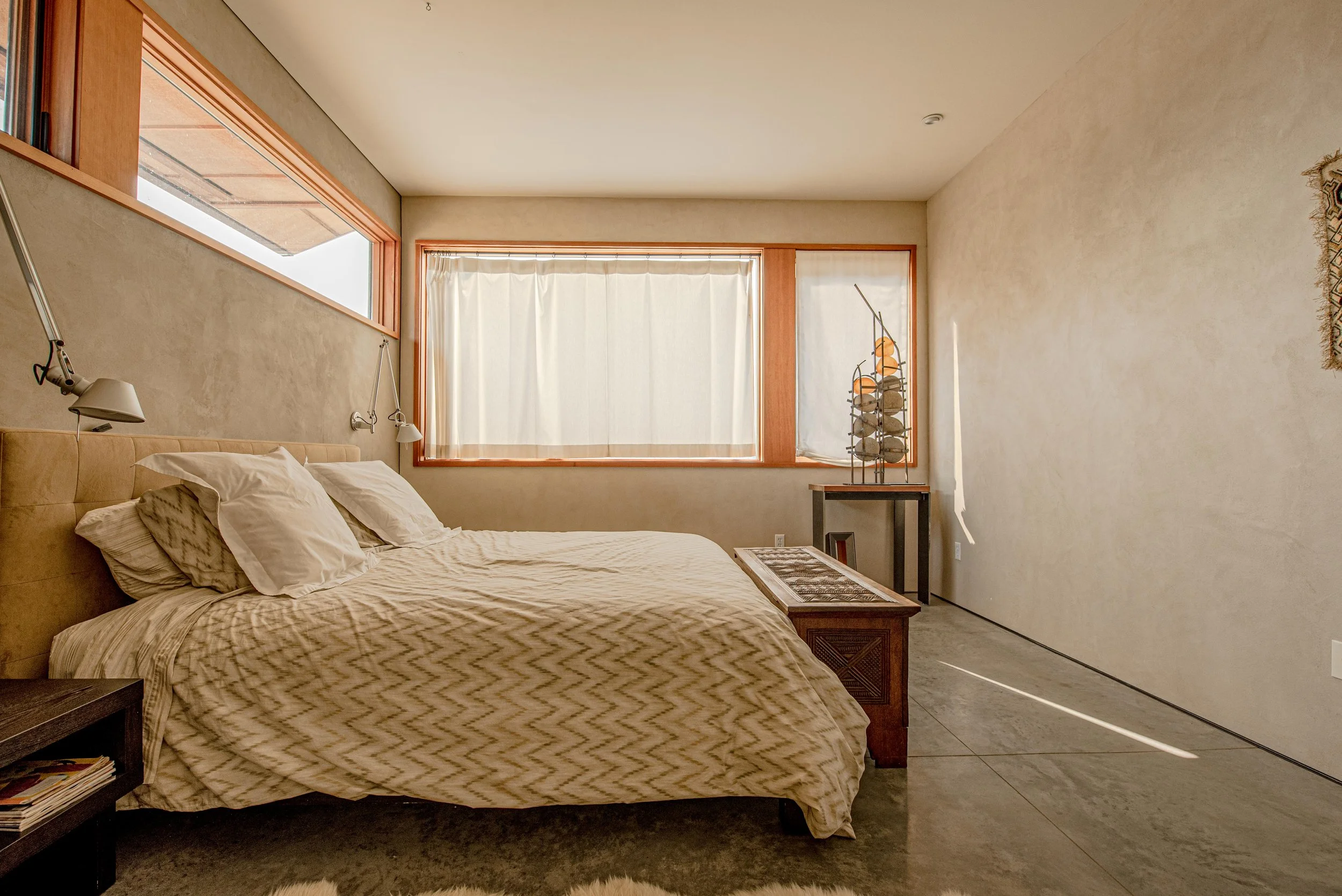 A bedroom with a bed featuring tan and beige patterned bedding and multiple pillows. There is a slit window with beige curtains, a side table with a lamp, and a decorative metal sculpture resembling a sailboat on a table. The room has a minimalist ae