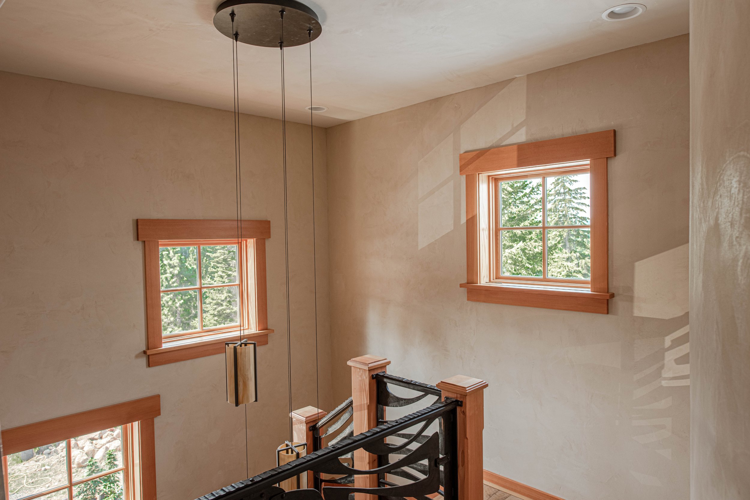 A room with beige walls and wooden window frames, showing two windows letting in sunlight and a view of green trees outside. There's a black metal railing at the bottom of the image, possibly part of a staircase, and a modern hanging light fixture ab