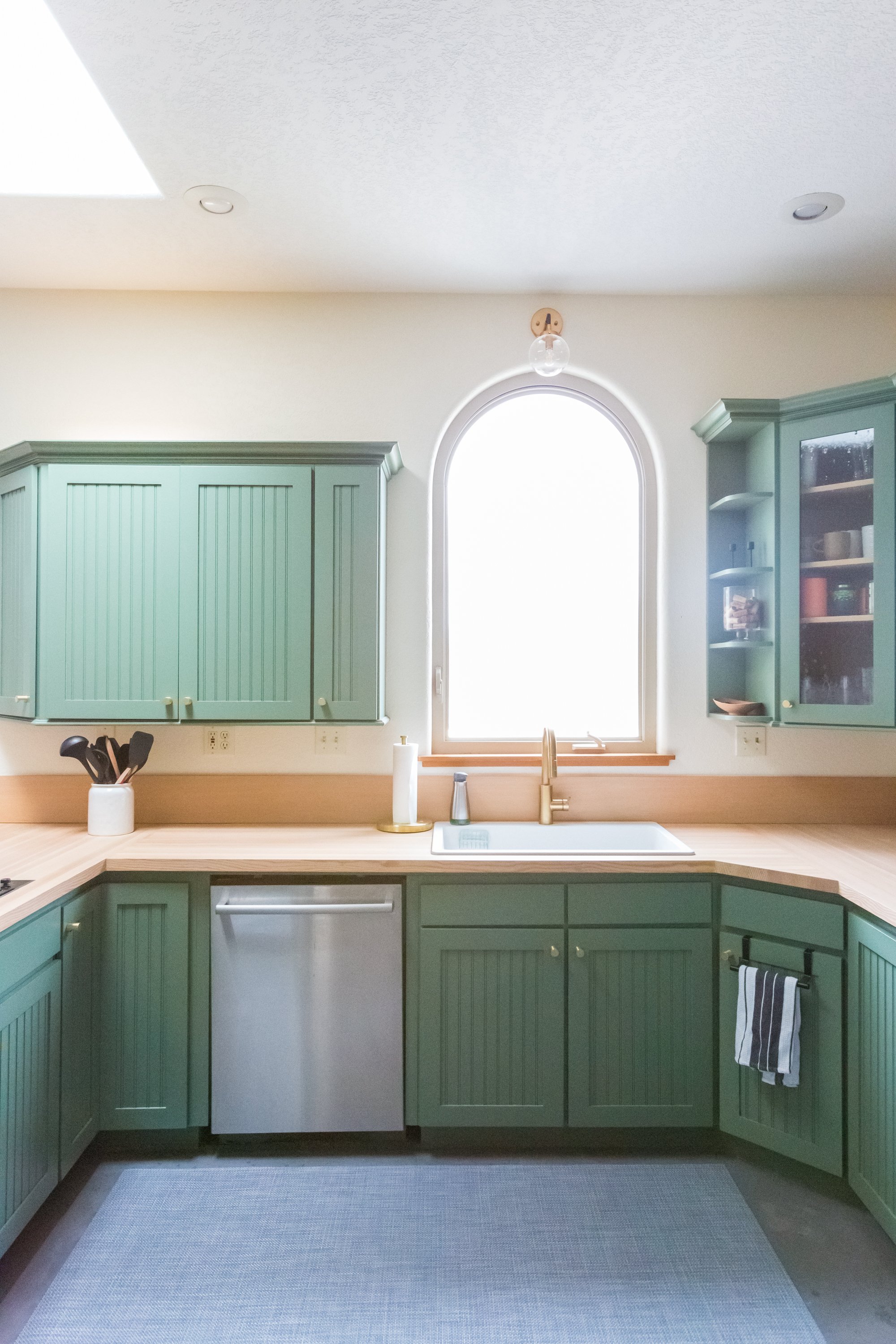 A modern kitchen with green cabinets, a white countertop, a stainless steel dishwasher, a white sink with a gold faucet, and a large arched window. There are black kitchen utensils in a white container and a striped towel hanging on the side.