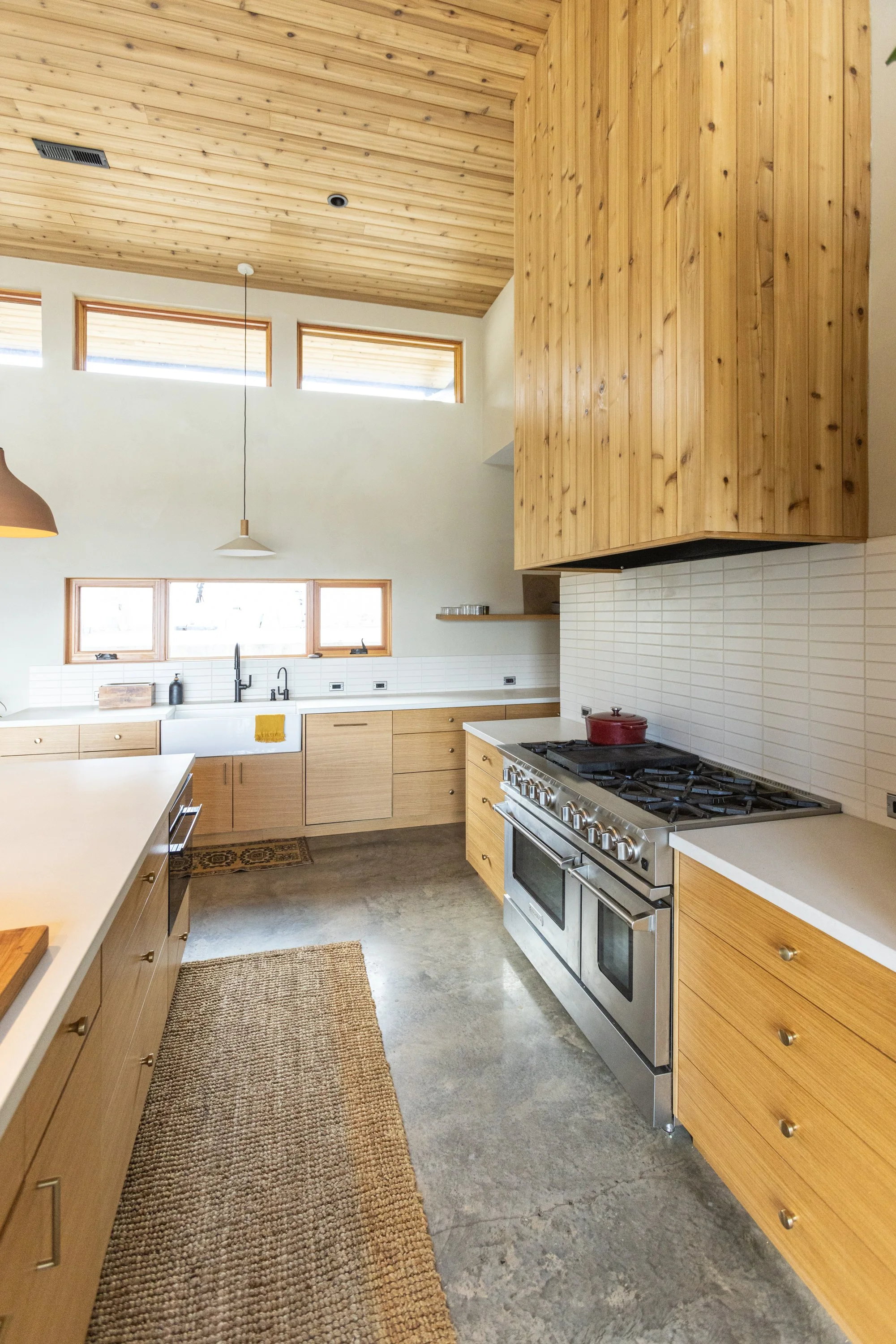 A kitchen with wooden cabinets, a stainless steel stove, and a rustic rug on a concrete floor.