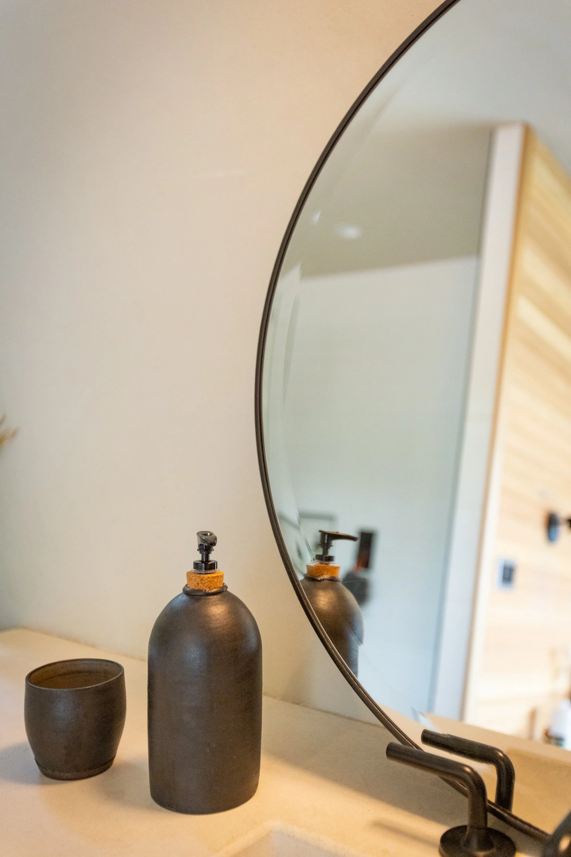 A bathroom countertop with a black soap dispenser and cup, a large oval mirror, and a window with wooden blinds reflected in the mirror.