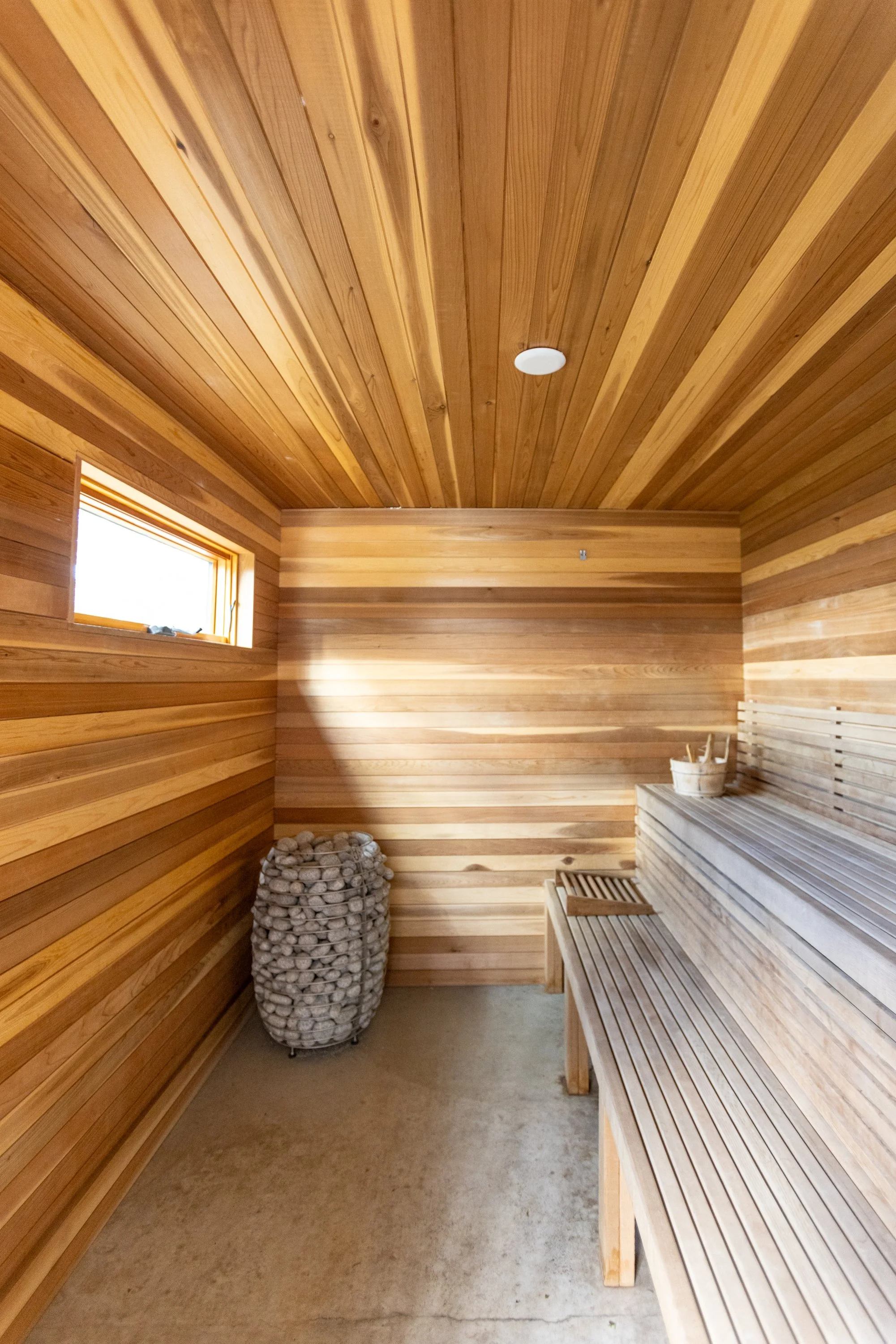 Wood-paneled sauna room with a wooden bench, a bucket of stones, and a container of sauna utensils. The room has a small window.