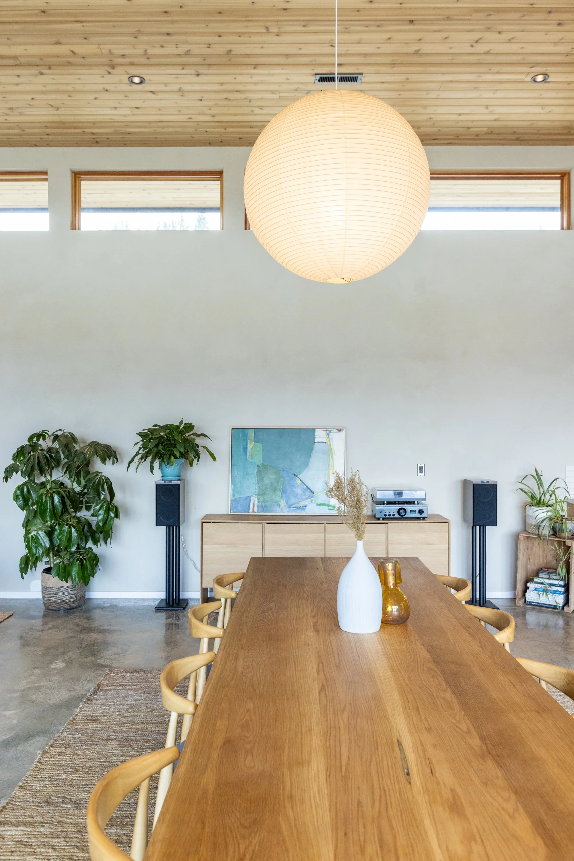 A dining room with a large wooden table, surrounded by wooden chairs, and a white vase with dried flowers on the table. The back wall has a wooden cabinet with artwork, potted plants, and a stereo system. A paper lantern hangs from the ceiling, and t
