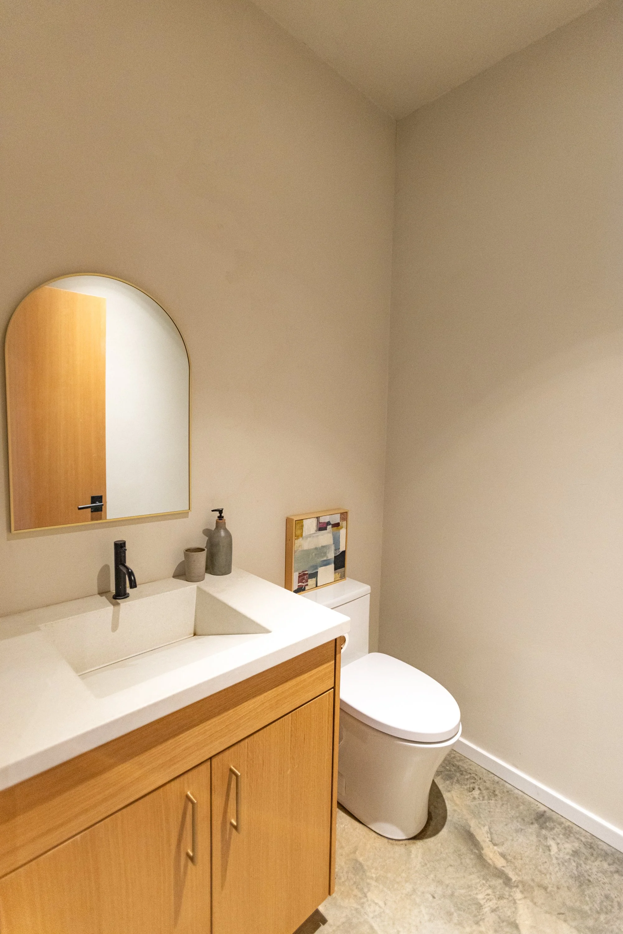 A bathroom with a wooden vanity, a white countertop, and a black faucet. Decorative objects and a framed art piece are on the counter. A round mirror hangs above the sink. There is a white toilet next to the vanity, and the walls are painted beige.