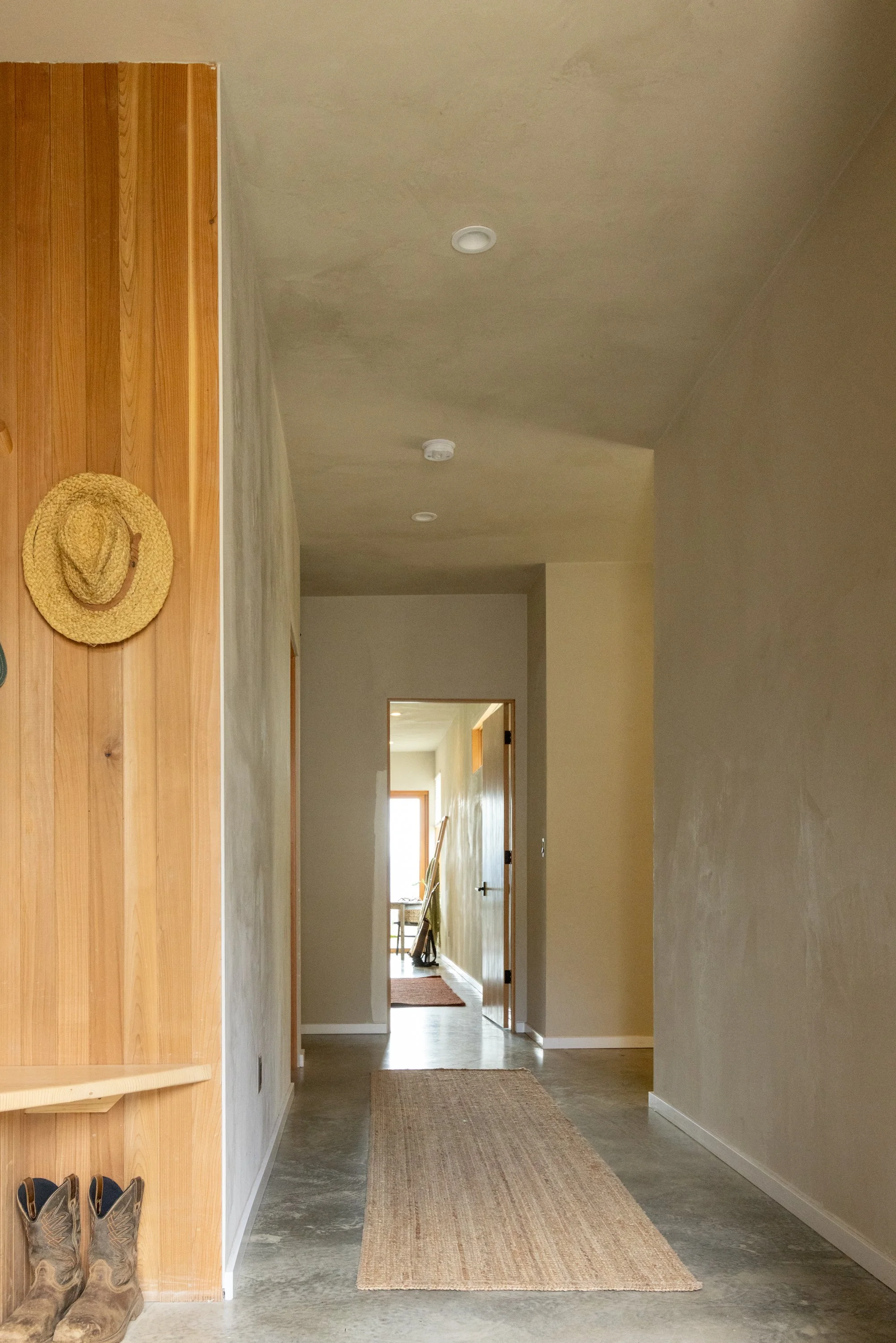 A hallway in a house with beige walls and a textured ceiling. A natural fiber rug runs along the concrete floor. To the left, there is a wooden wall with a straw hat hanging on it and a pair of cowboy boots on the floor. At the end of the hallway, th