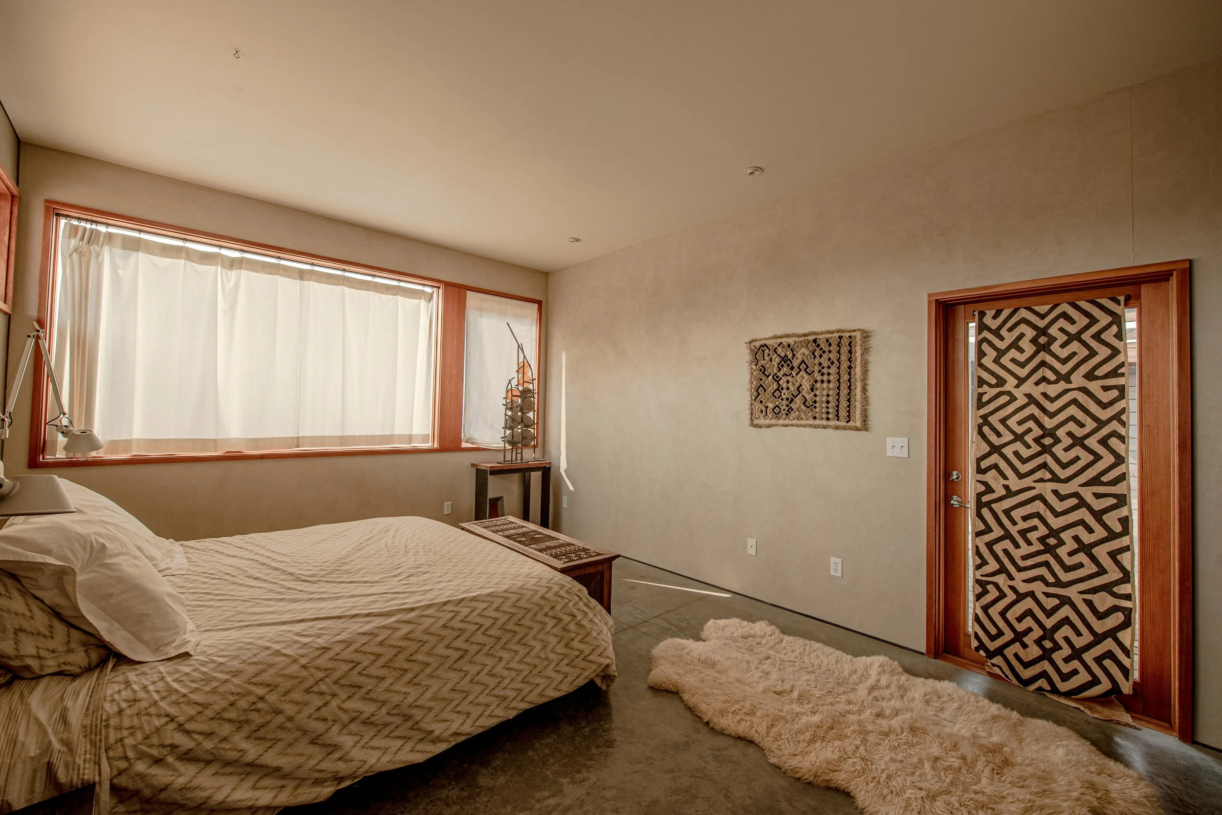 A bedroom with a bed, large window with white curtains, a side table with decorative items, a wall hanging, and a door with a patterned fabric covering.