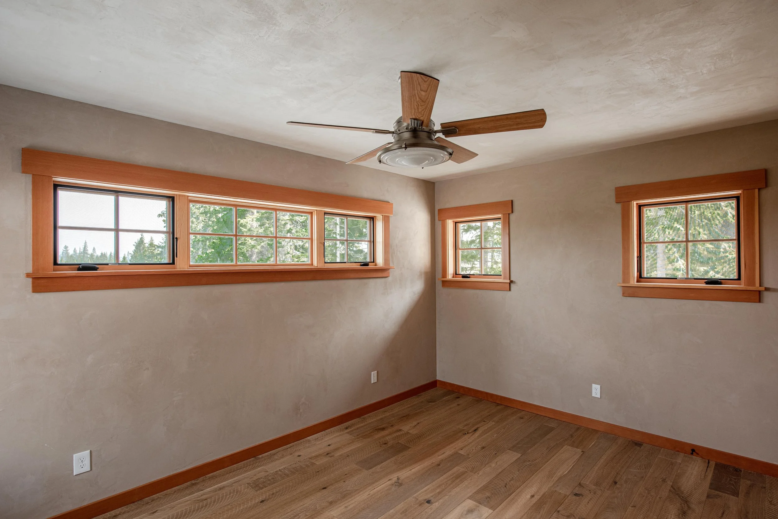 Empty room with three rectangular windows and wooden trim, beige walls, wooden flooring, ceiling fan with wooden blades, and electrical outlets.
