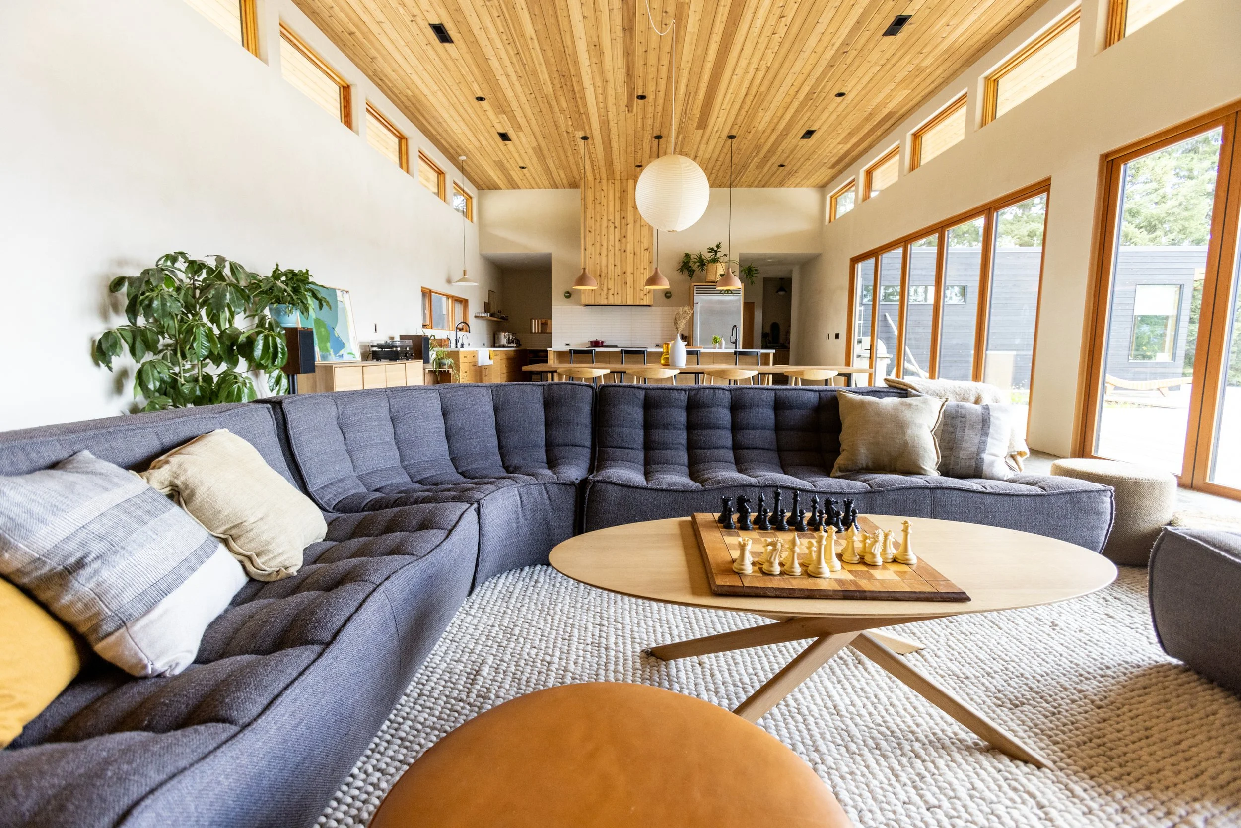Living room with a large dark sofa, a round wooden coffee table with a chessboard, and a cream-colored rug. There is a kitchen in the background with wooden cabinets, a white backsplash, and large windows letting in natural light.