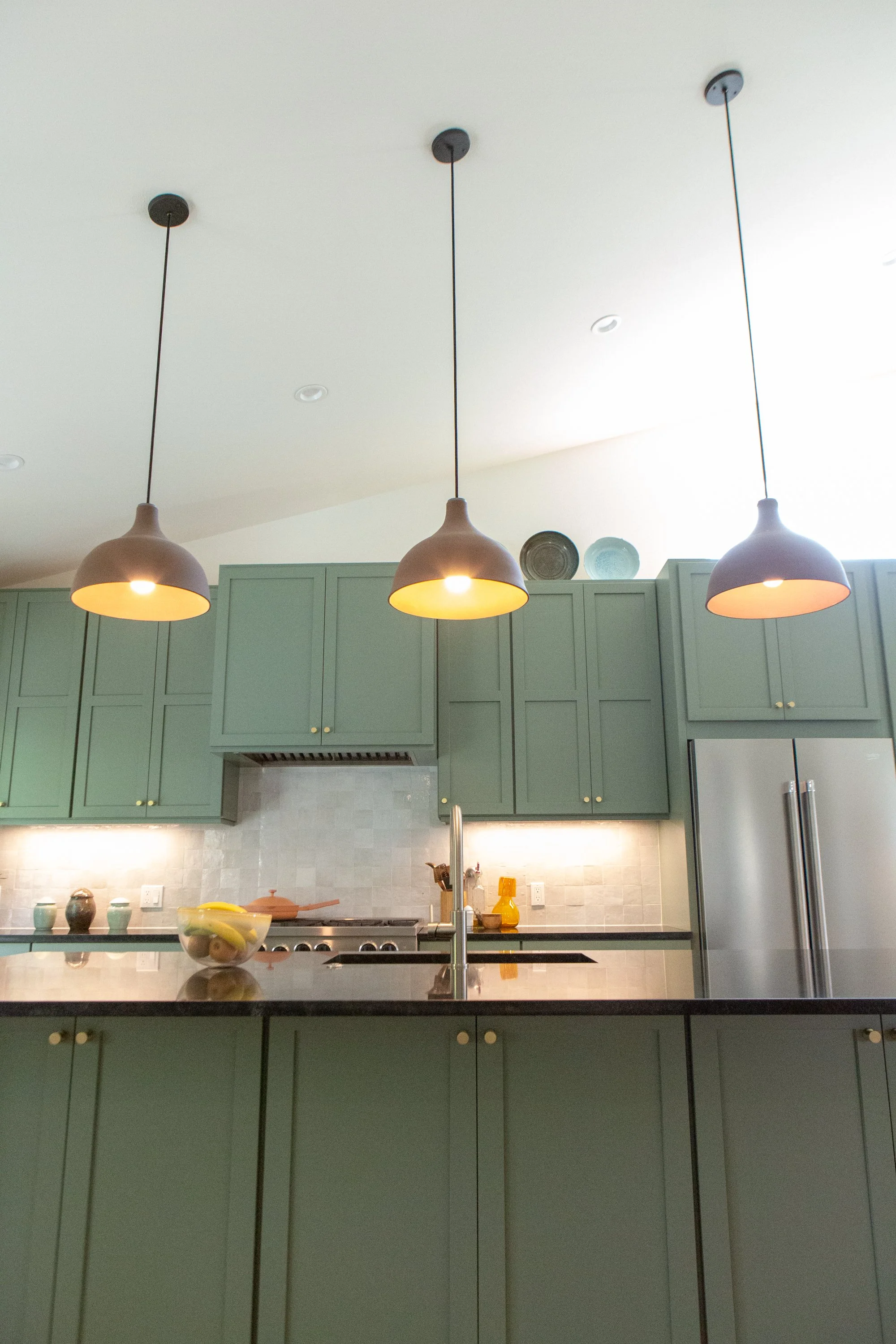 Modern kitchen with over head light fixtures, green cabinets, a bowl of fruit on the counter, and decorative plates on top of the cabinets