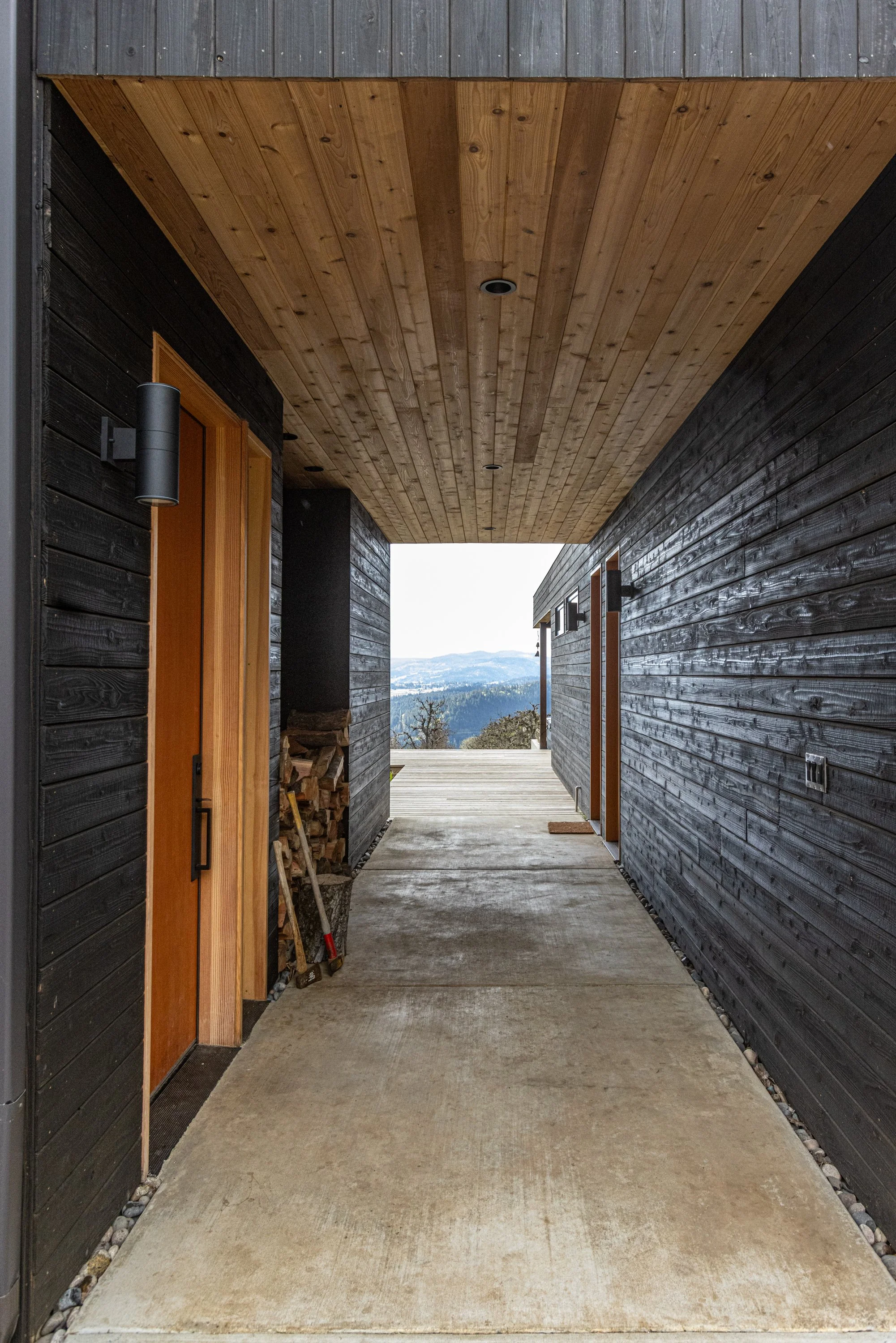 A covered outdoor corridor with a concrete floor, black wooden walls, and a light wooden ceiling. There are doorways on the left and right, and in the background, mountain scenery can be seen.
