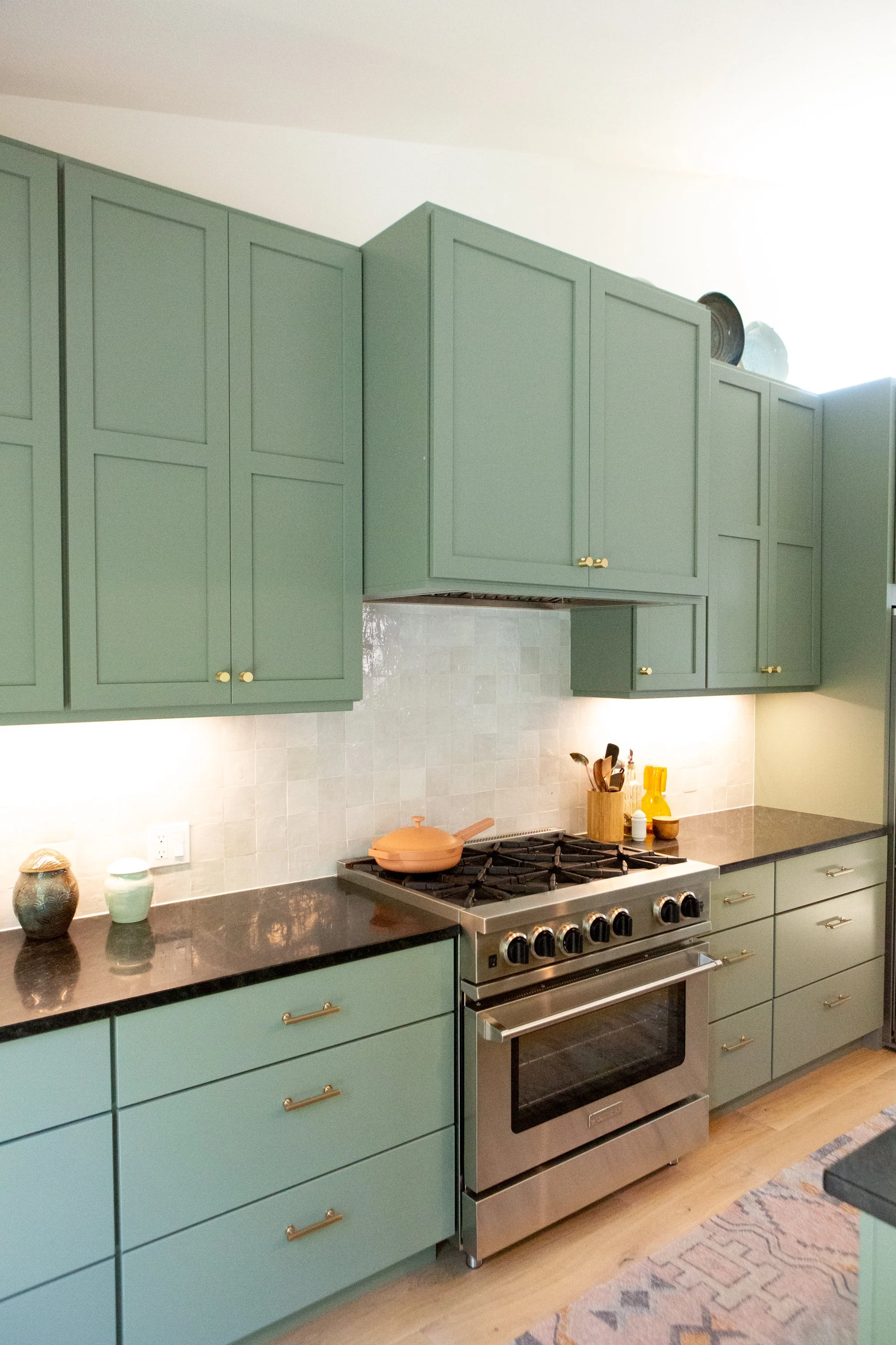 Green kitchen cabinets above a black countertop with vases and a ceramic dish. Stainless steel gas stove with oven and hardwood floor. Patterned rug in front of the stove.