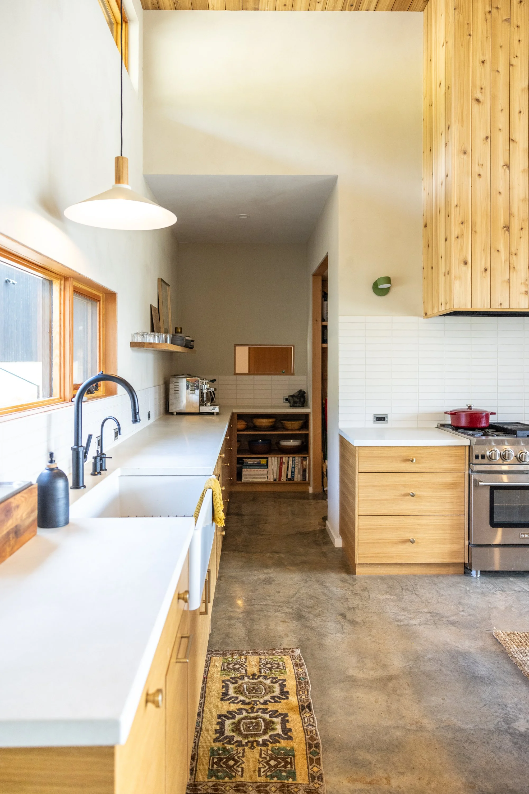 A modern kitchen with wooden cabinets, a white countertop, and stainless steel appliances. There are windows on the left side, and a decorative rug on the polished concrete floor.