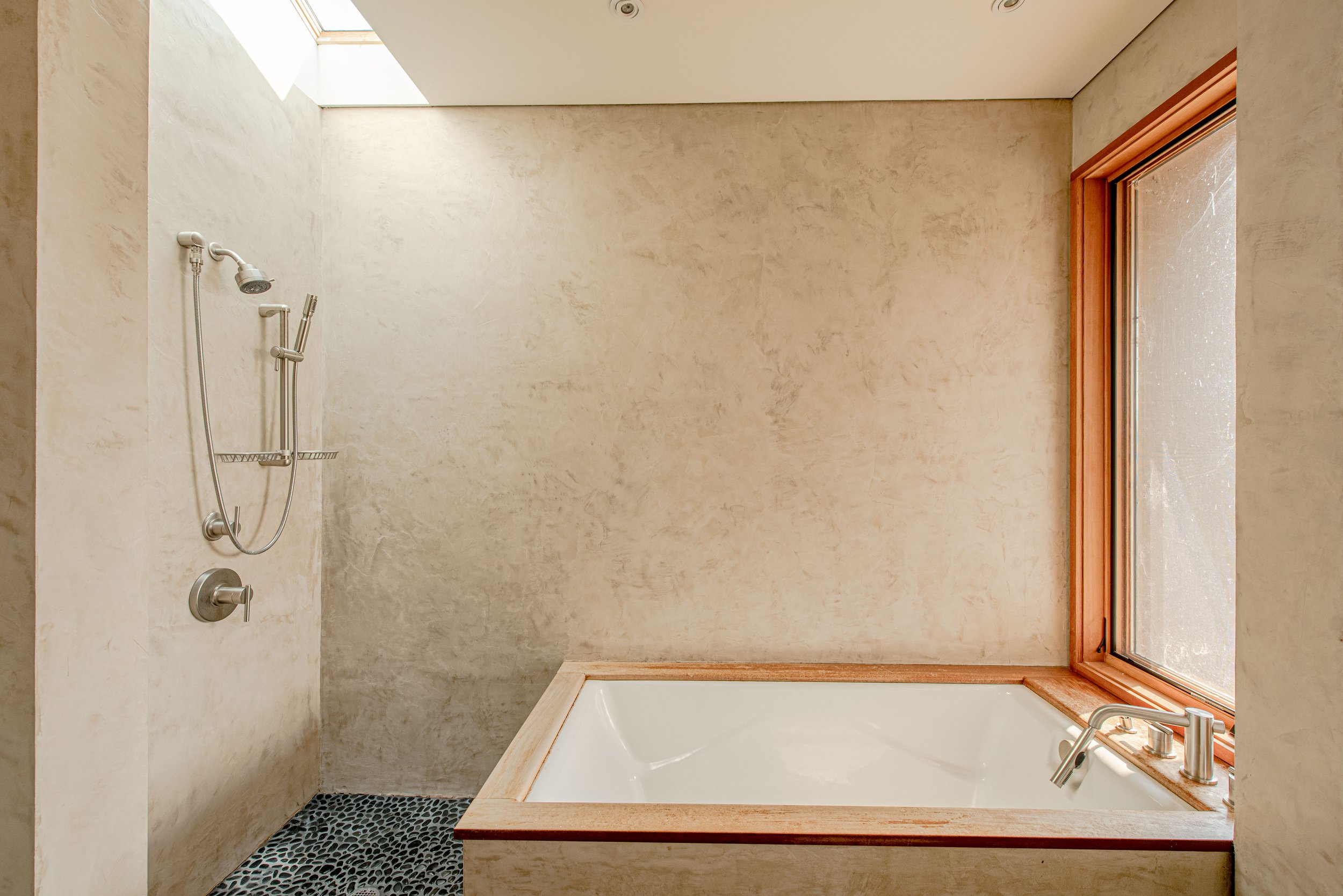 A modern bathroom with a large soaking tub, beige textured walls, a corner shower with two showerheads, a small patterned black floor mat, and a large window with a wooden frame.
