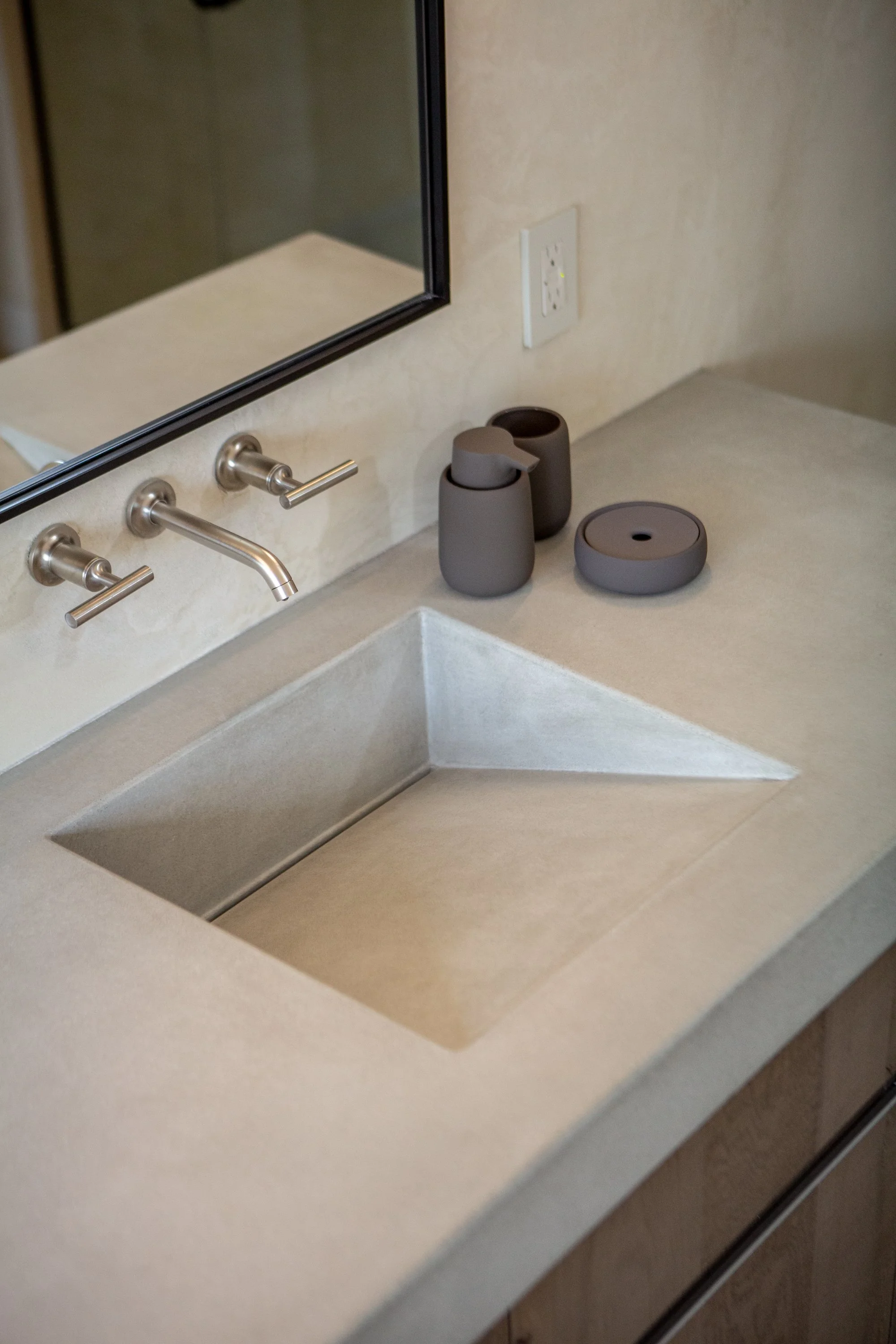 A bathroom sink with a modern faucet, a mirror, and two dark gray containers on the countertop. One container is a soap dispenser, and the other is a cup. There is also a round gray container with a hole on top. An electrical outlet is visible above 