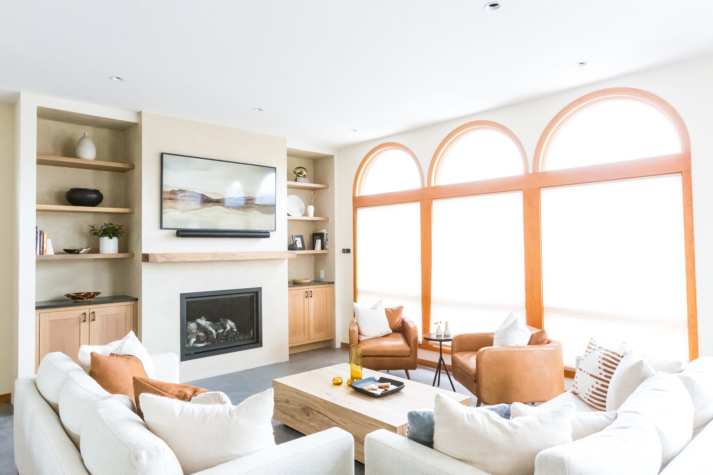Living room with a white sofa, brown leather armchairs, a wooden coffee table, a wall-mounted flat-screen TV, and large arched windows with orange trim, decorated with vases and framed pictures.