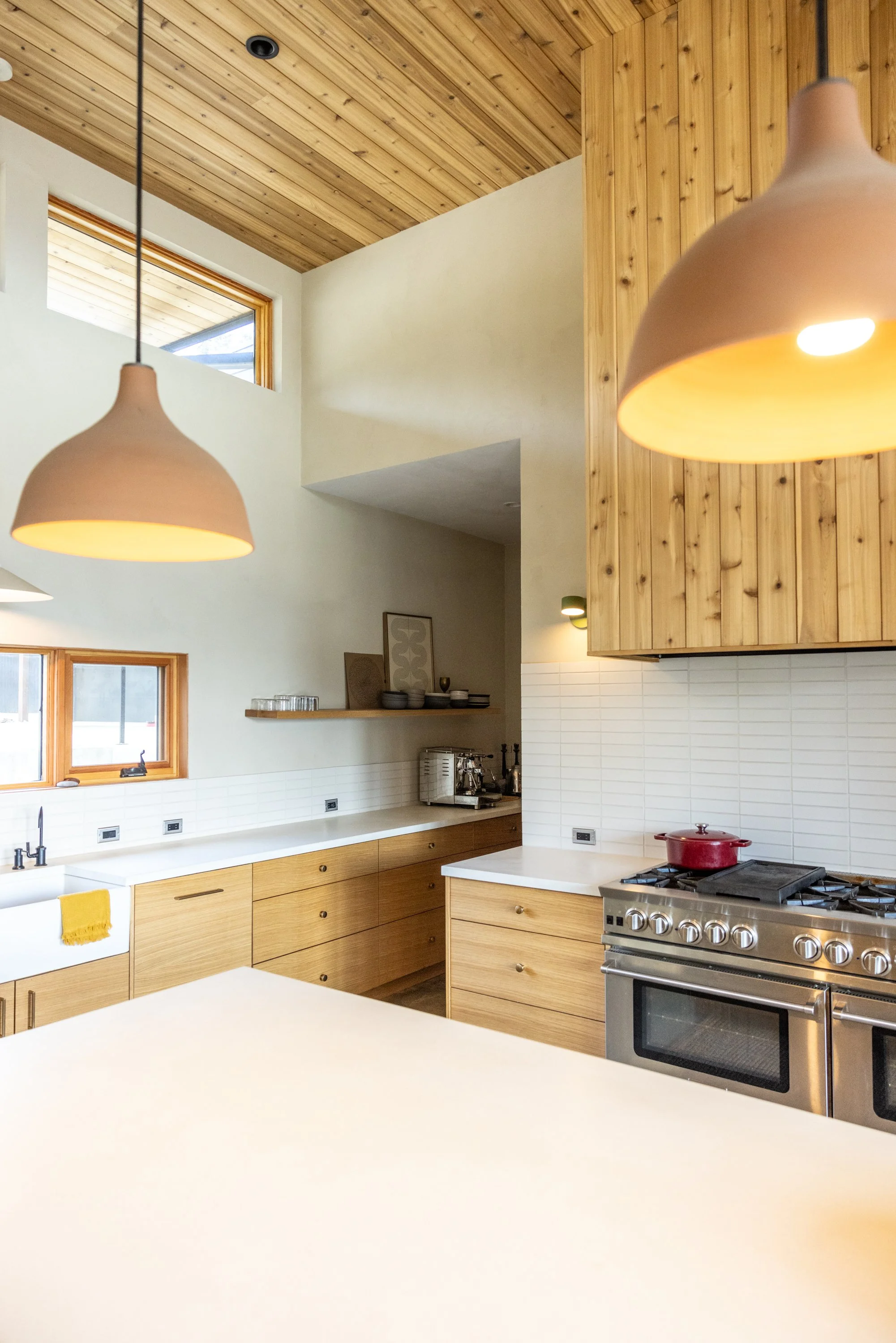 Modern kitchen with wood cabinetry, white countertops, stainless steel stove with red pot, open shelving with dishes and bowls, and large wooden ceiling with two hanging pendant lights.
