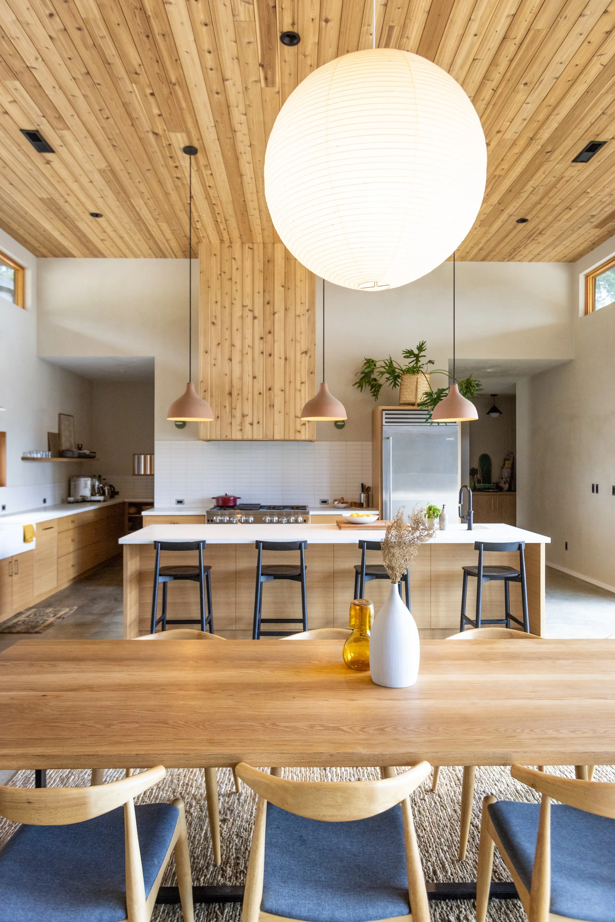 Interior view of a modern kitchen with a large paper lantern ceiling light, wooden ceiling and cabinetry, white countertop, and black bar stools at an island. Decor includes vases with dried flowers and a green plant.