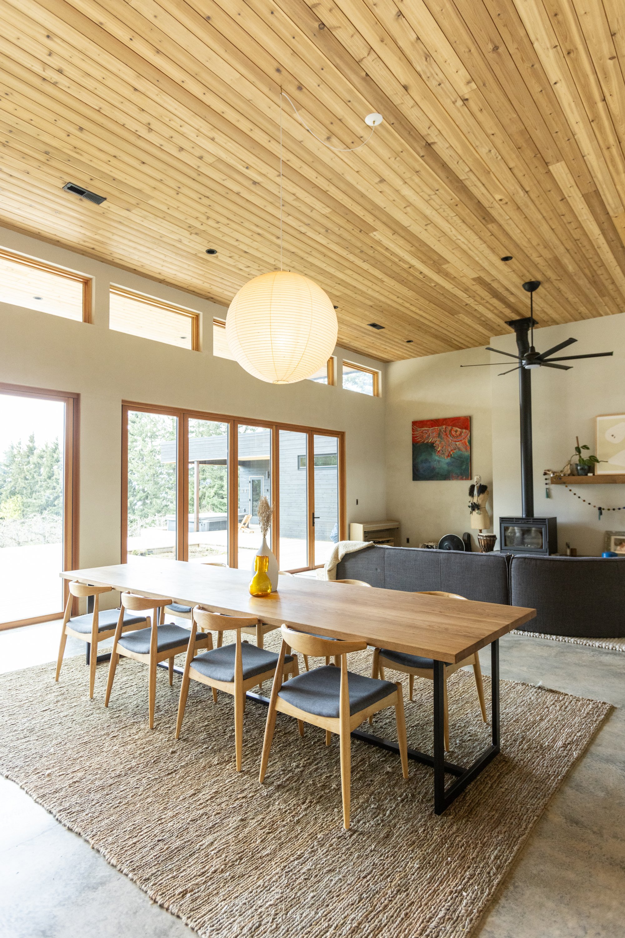 Open living and dining area with a wooden ceiling, large windows, a dining table with chairs, a rug, a sofa, a wall-mounted painting, a fireplace, and a ceiling fan.