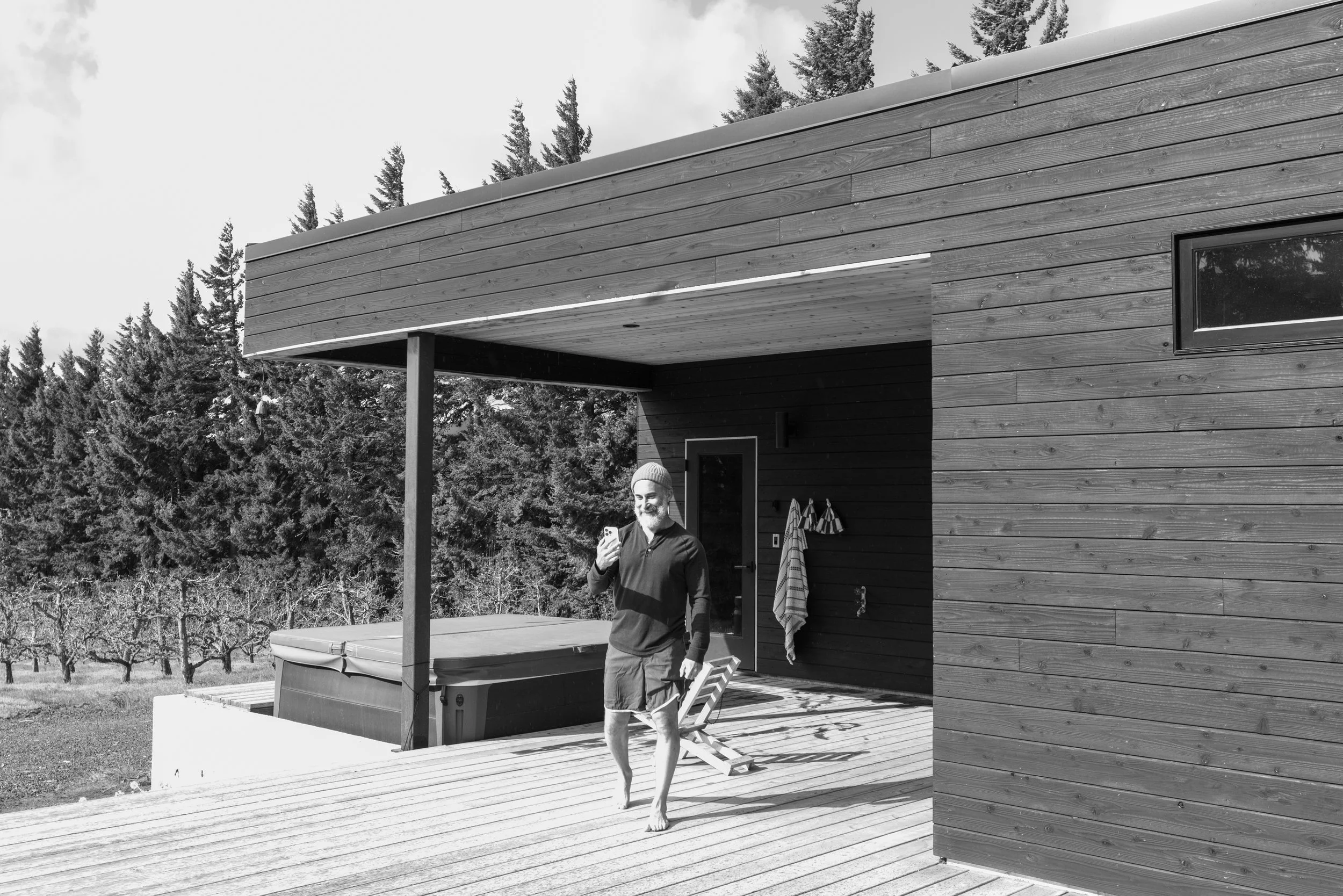 A man with a beard and cap is standing on a wooden deck, looking at his phone, outside a modern house with wood siding and a small window, surrounded by trees.