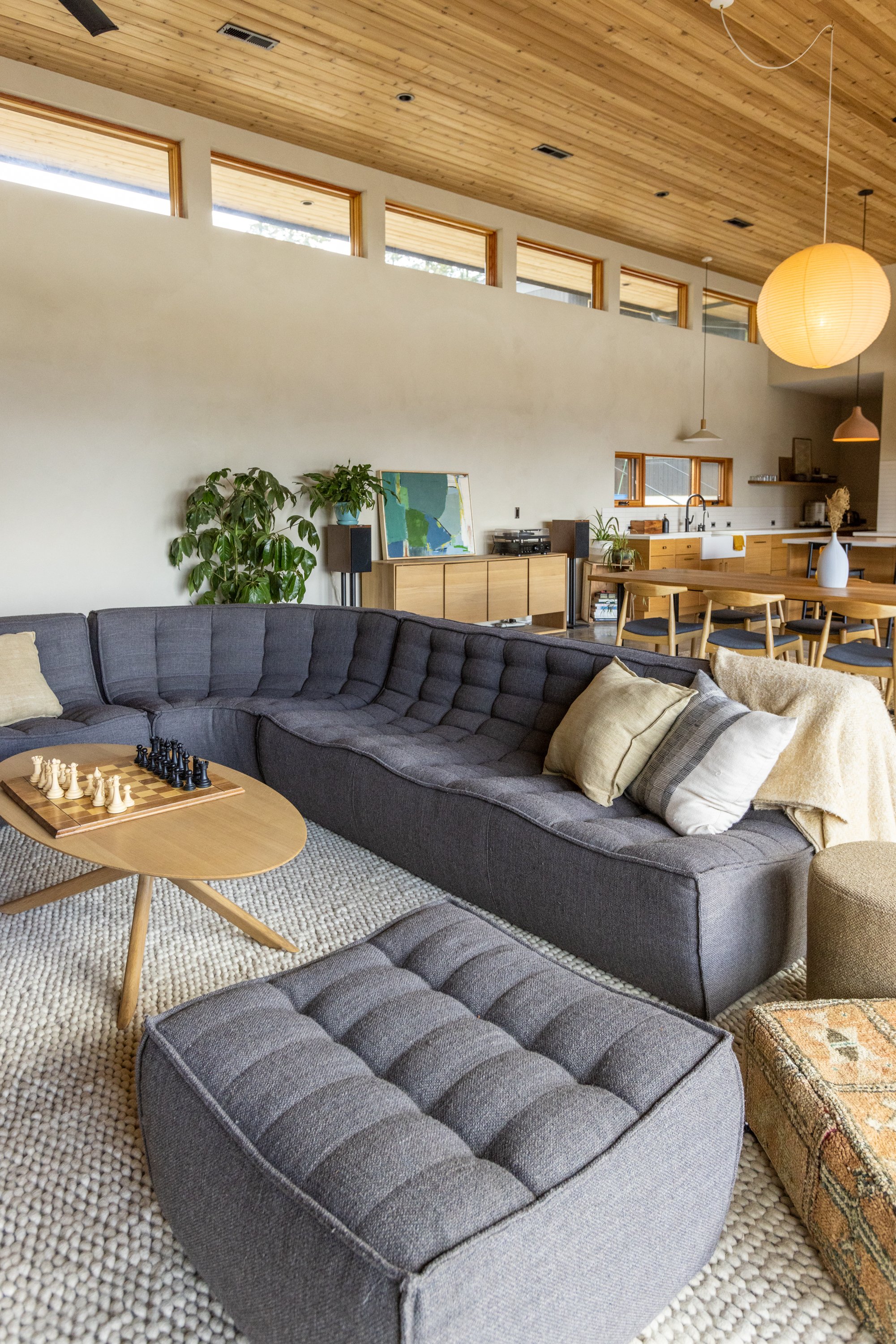 Cozy living room with dark gray sectional sofa, beige pillows, round wooden coffee table with chessboard, beige textured rug, houseplants, and wooden ceiling with natural light from rectangular windows.