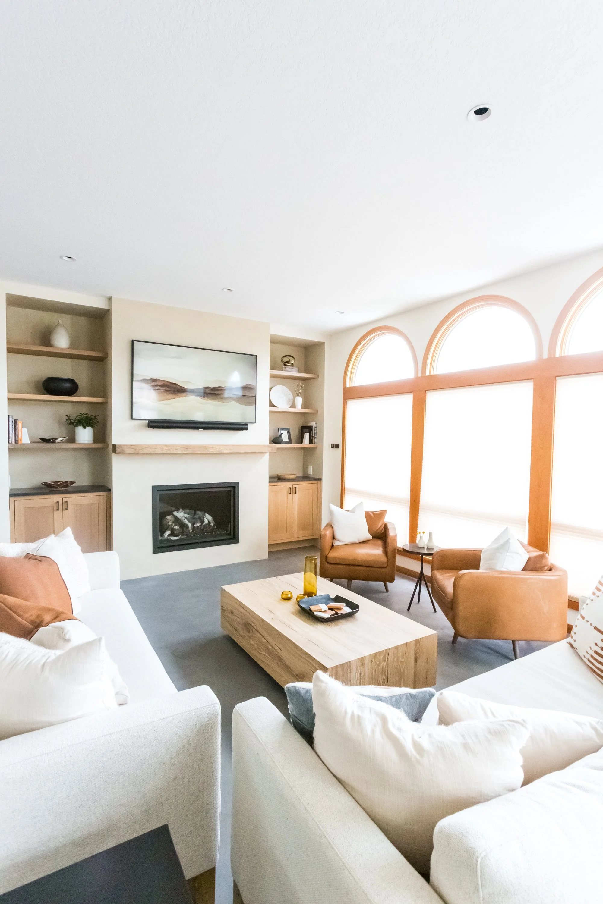A living room with white sofas, leather armchairs, a wooden coffee table, built-in shelves with decorative items, a fireplace, and a wall-mounted TV. Large arched windows allow natural light to fill the space.