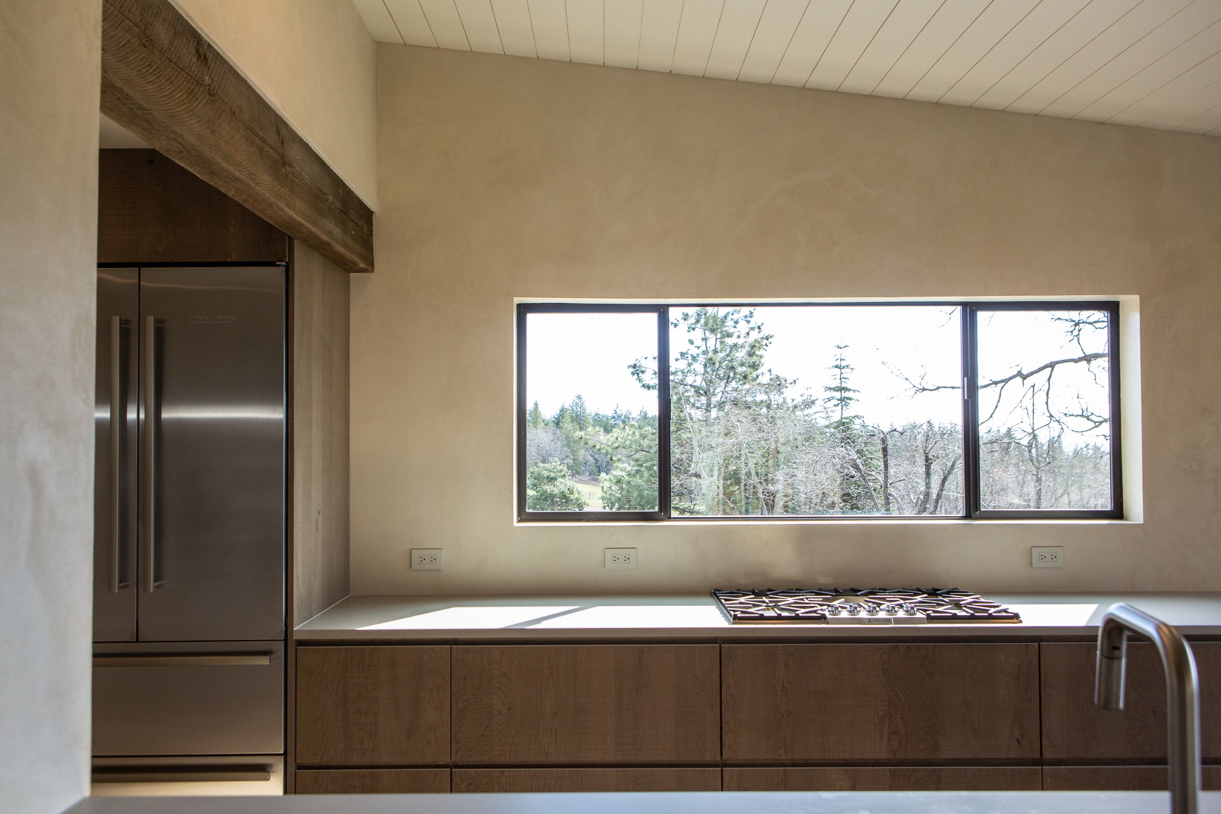 Modern kitchen with large window showing trees outside, a stainless steel refrigerator, and a gas stove on a wooden countertop.