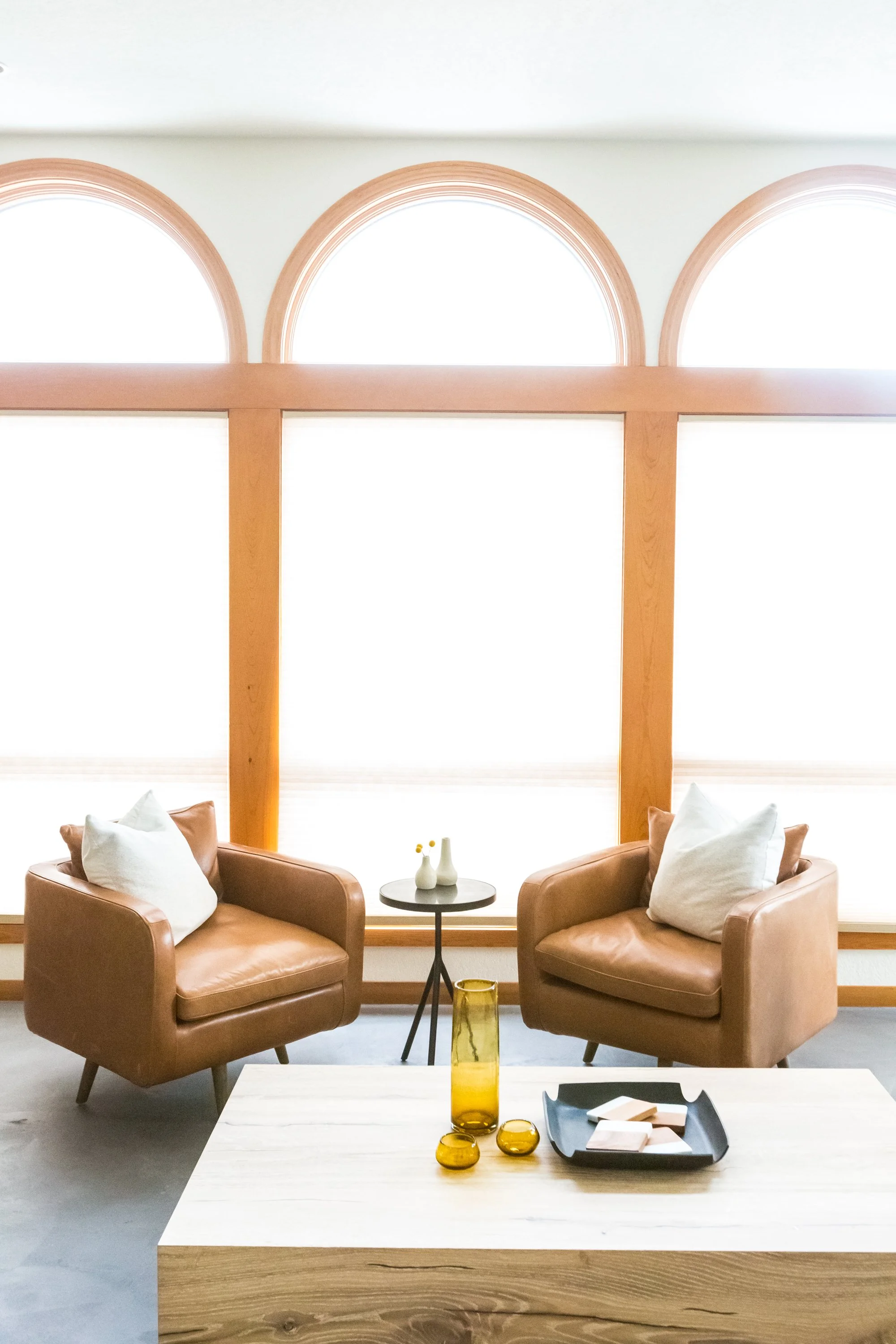 Living room with two brown leather armchairs with white pillows, a small black side table with white vases, a large wooden coffee table with decorative items, and large arched windows with wooden frames letting in bright natural light.