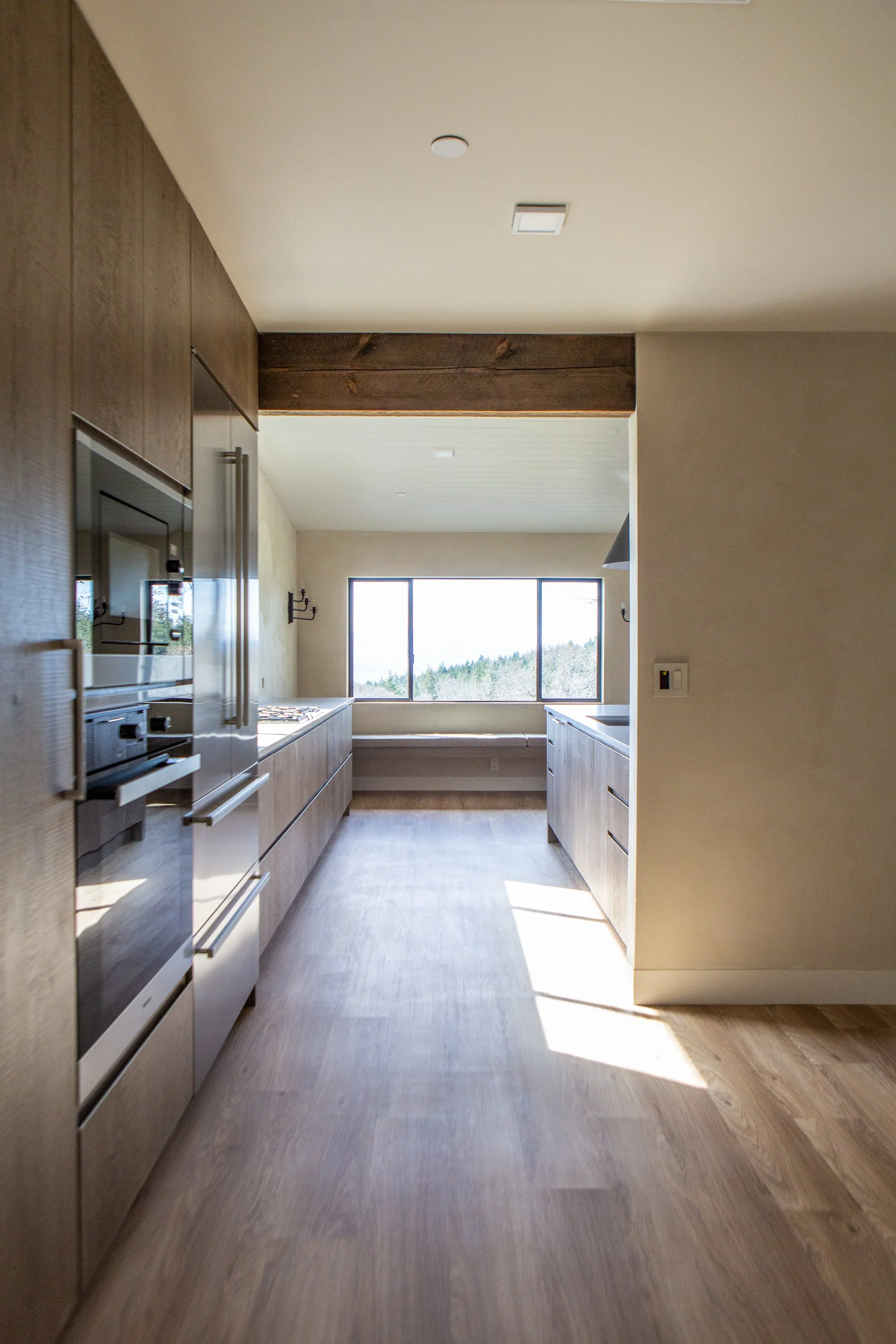 Kitchen with wooden cabinets, stainless steel appliances, a large window showing an outdoor view, and hardwood flooring, with sunlight casting shadows.