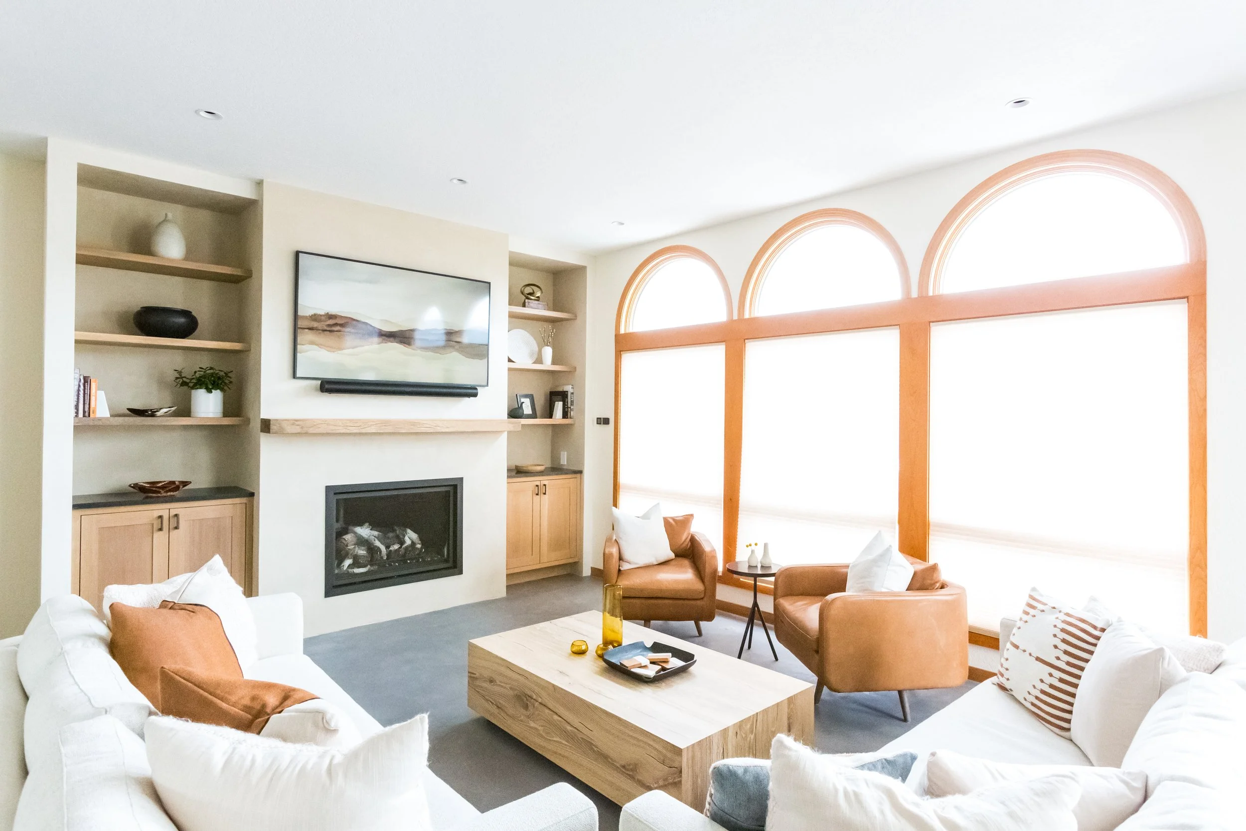 A modern living room with white sofas, leather armchairs, and wooden accents, featuring large arched windows letting in bright sunlight.