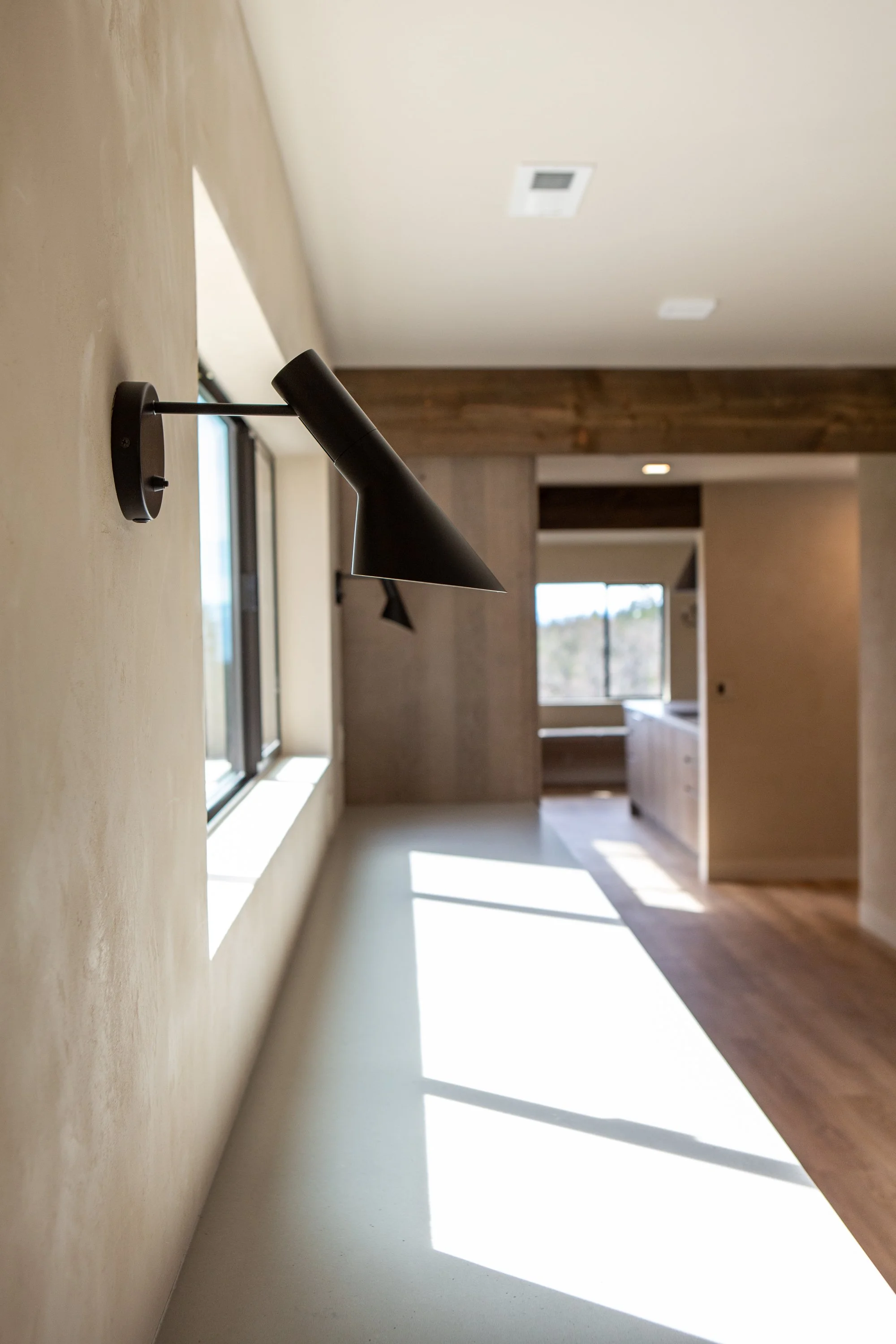 Interior of a modern house with a black wall-mounted reading light, cream-colored wall, windows, and natural light casting shadows on the light-colored floor.