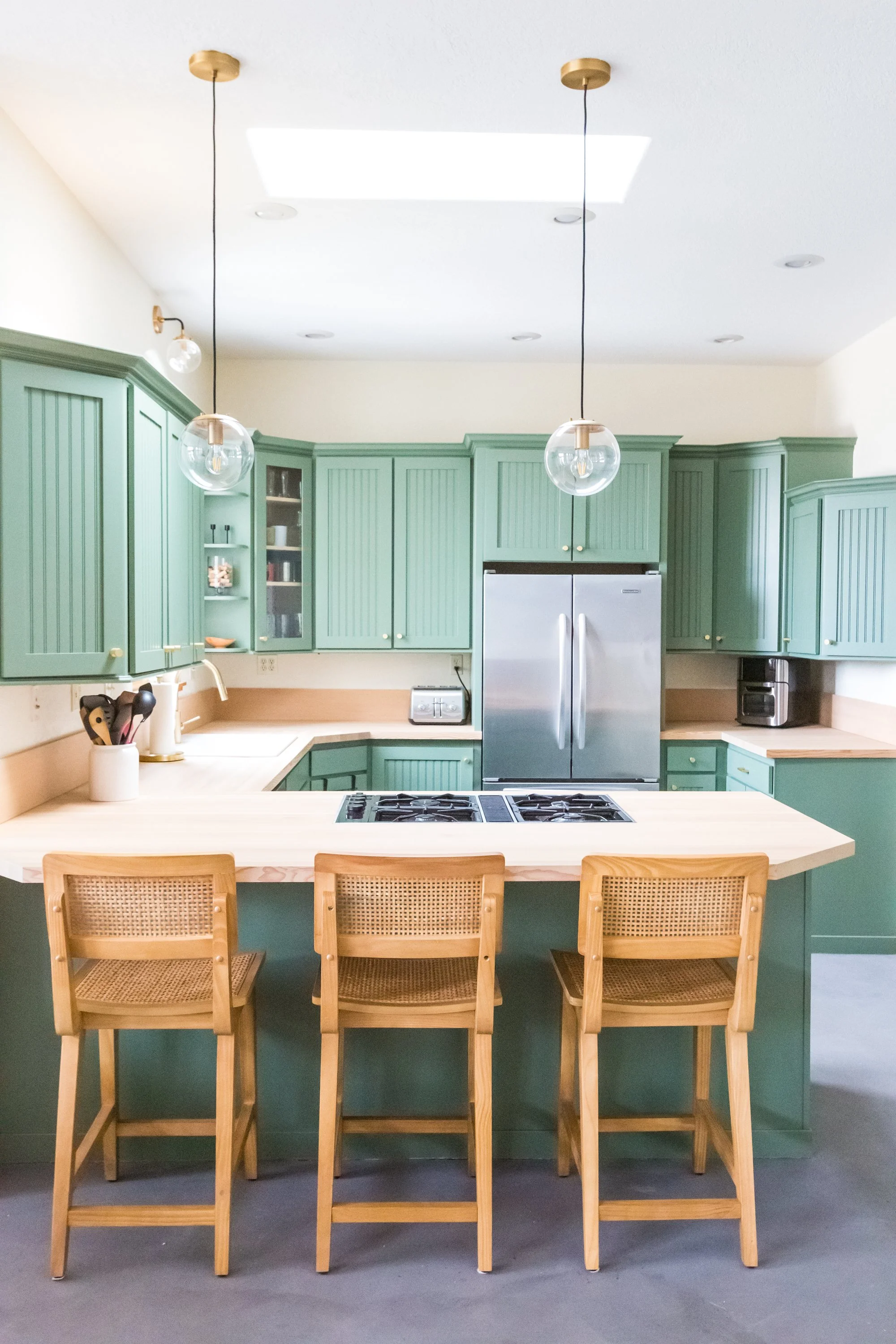 A modern kitchen with green cabinets, wood countertops, and a central island with four wooden chairs. There are two pendant lights hanging from the ceiling, a skylight, and various small appliances on the counters.