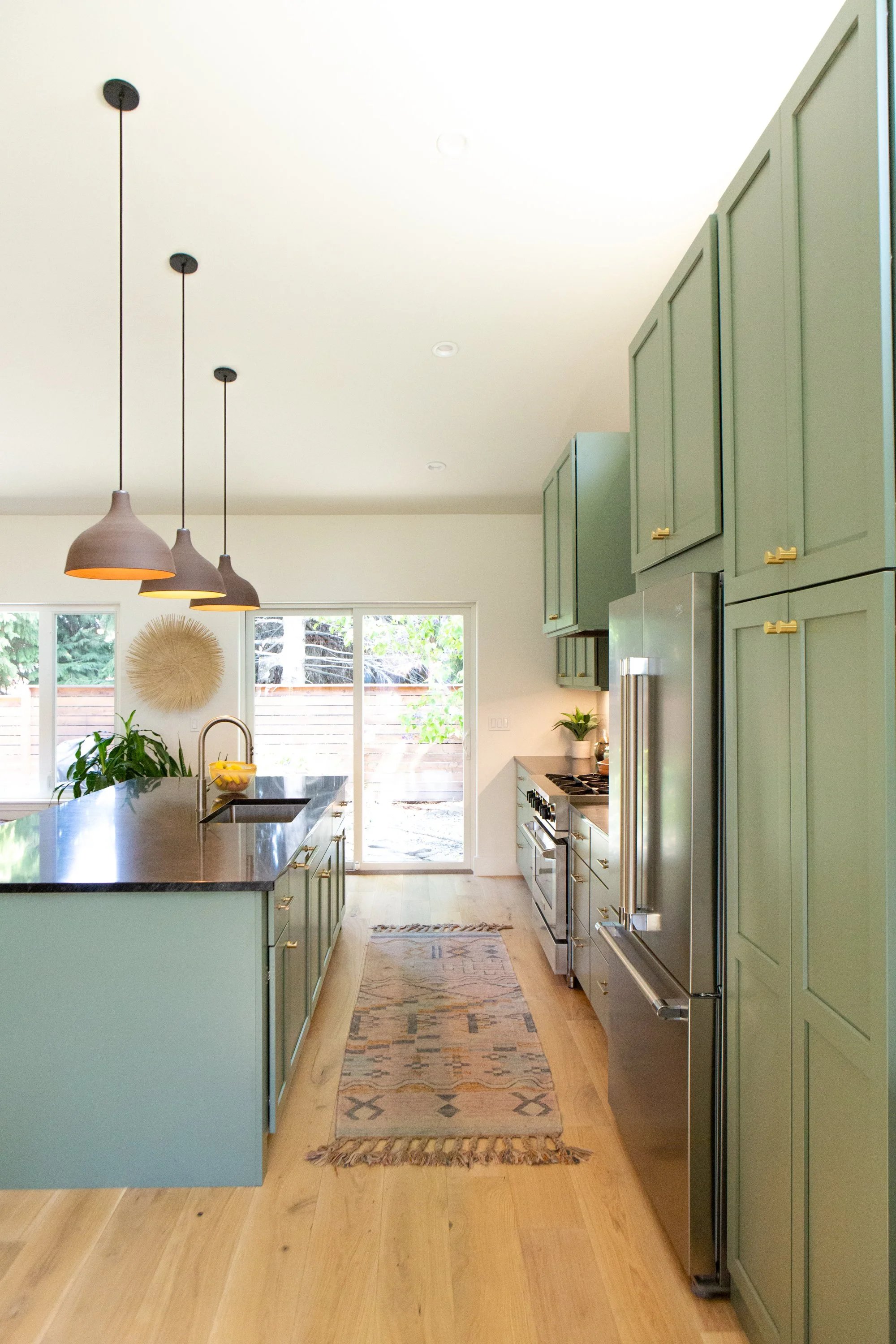 A modern kitchen with green cabinets, a black countertop island, three hanging pendant lights, a stainless steel refrigerator, and a window with a view of trees outside.