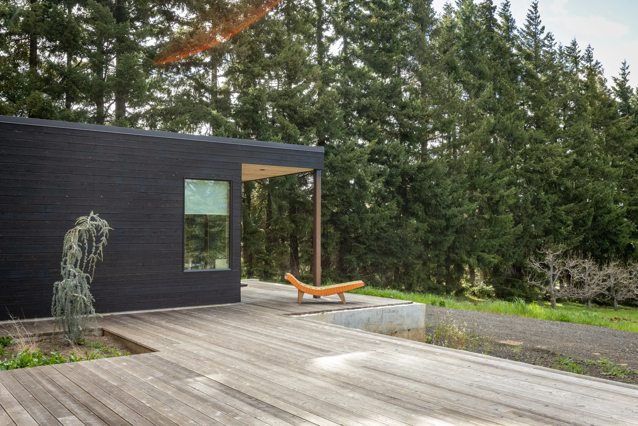 A modern black house with a wooden deck, surrounded by lush green trees, and an orange lounge chair on the deck.