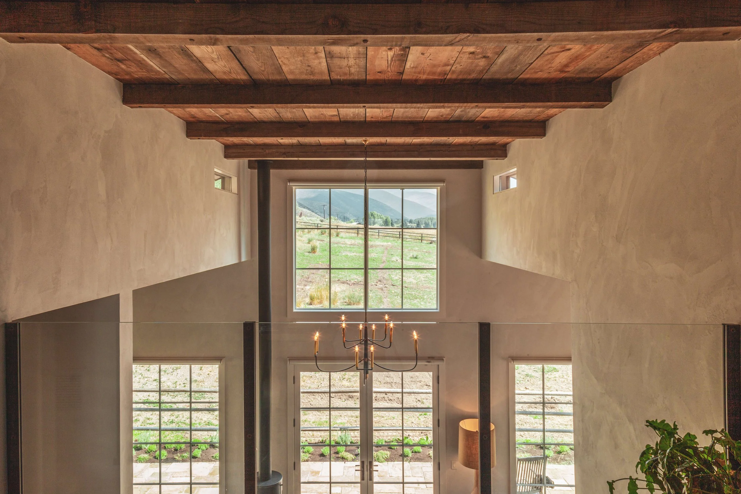 Interior view of a modern house with high ceilings, large windows showing outdoor landscape with mountains, hanging chandelier, and polished wooden beams.