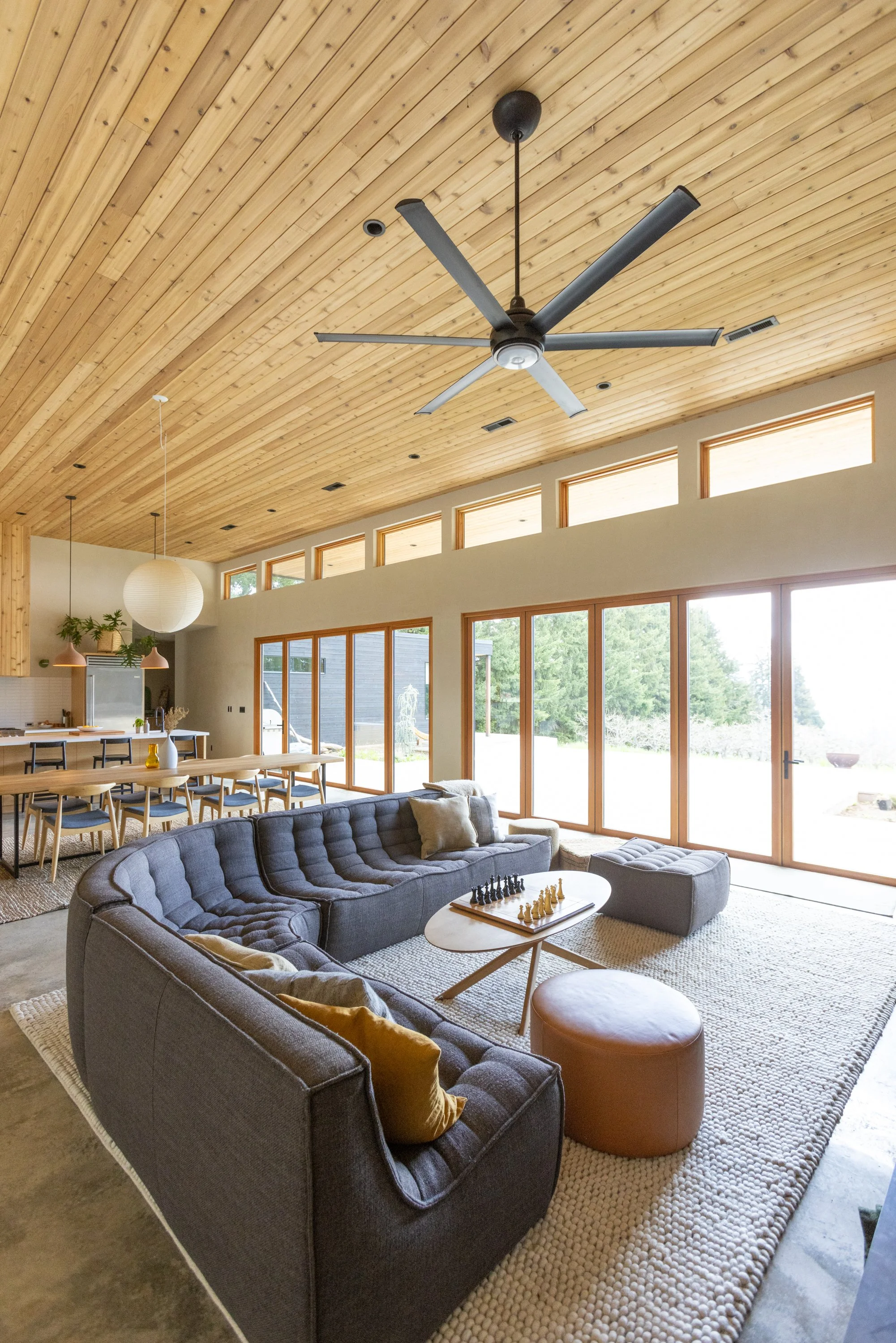 Living room with a curved gray sofa, a white round coffee table with a chessboard, and a beige stool. Large windows and glass doors provide a view of the outdoors, with a wooden ceiling and modern decor.