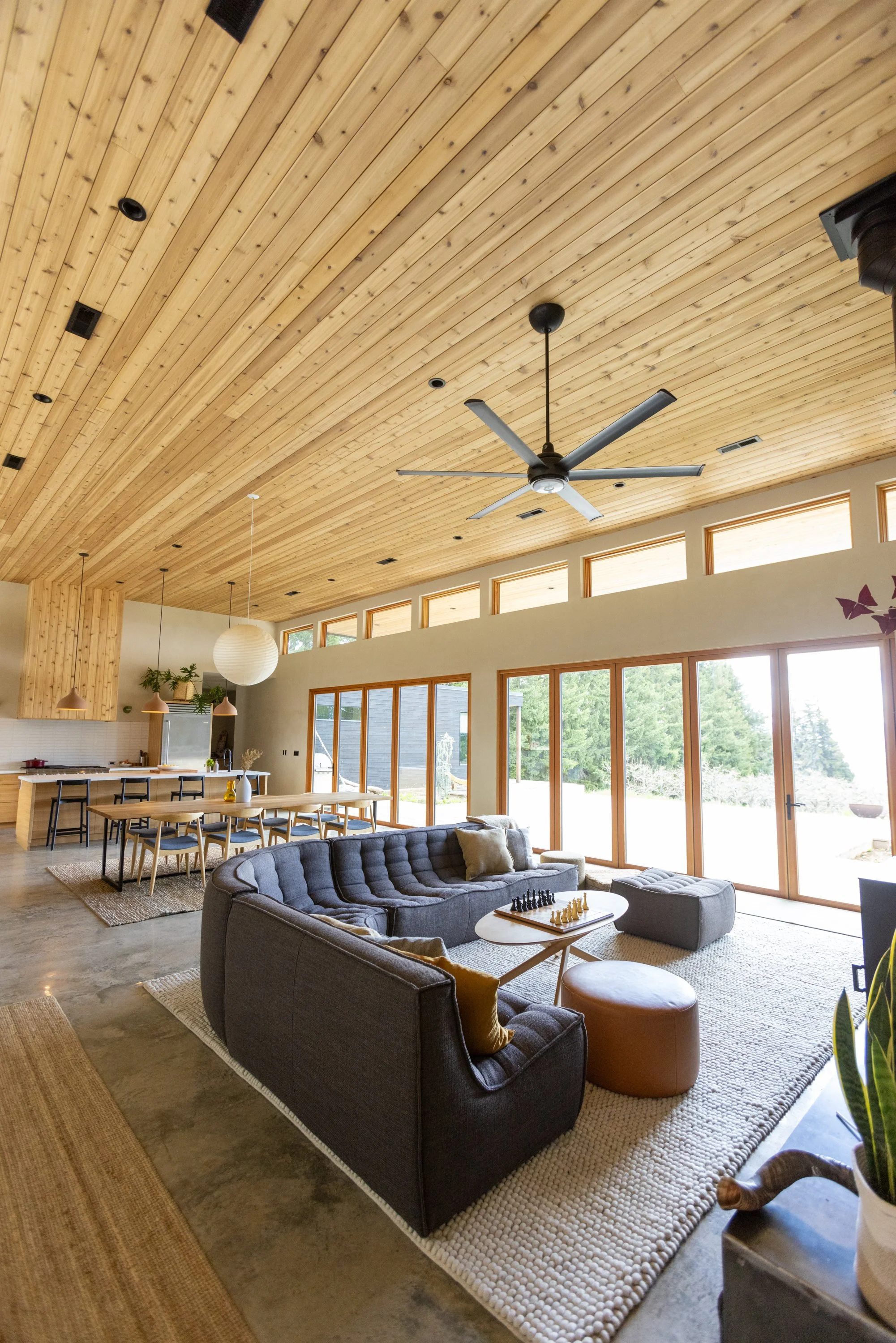 Living room with large windows, wooden vaulted ceiling, ceiling fan, and modern furniture including a gray sofa, coffee table with chess set, and a dining table with chairs.