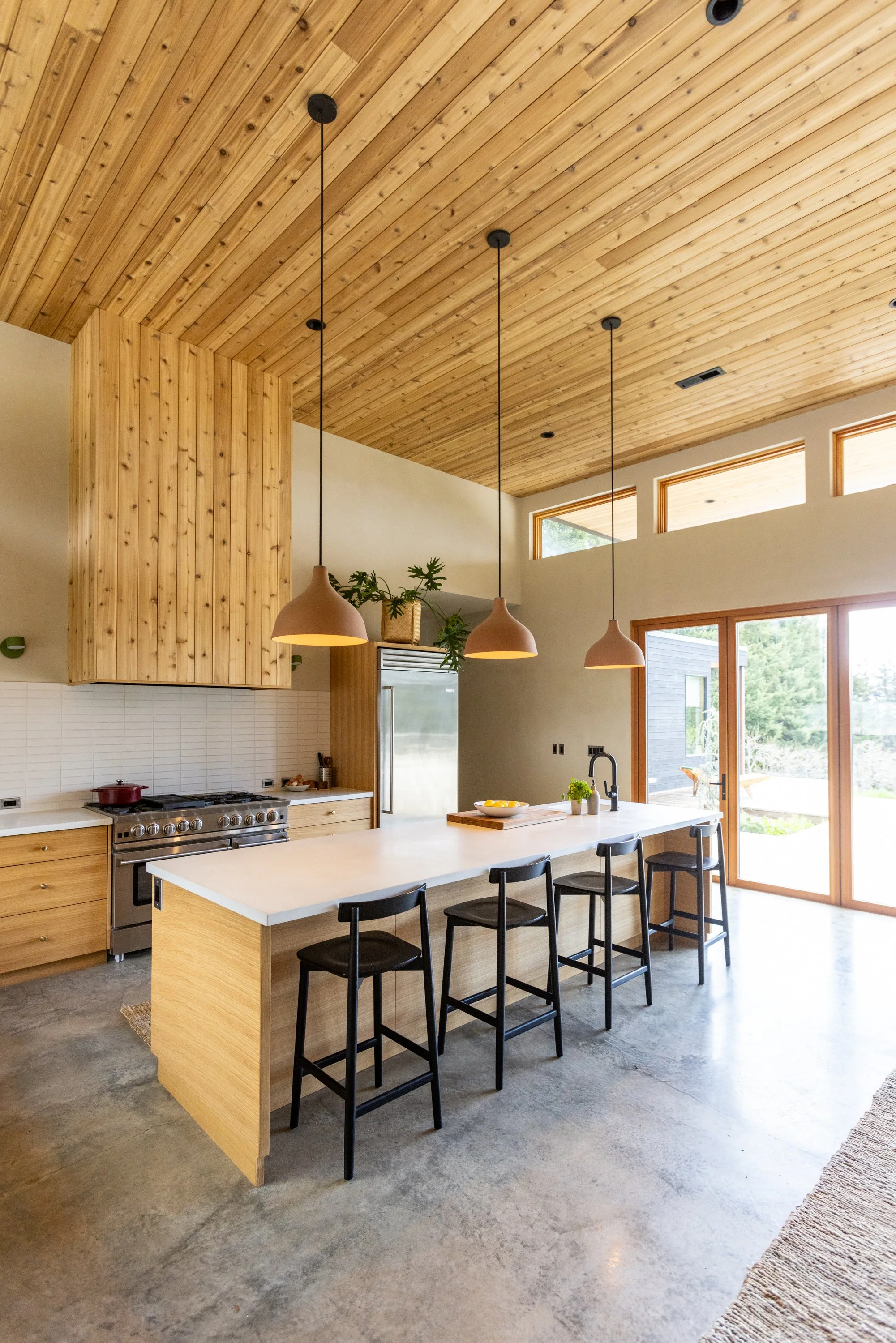 Kitchen with a large white island, black bar stools, wooden cabinets, pendant lights hanging from a wood-paneled ceiling, large windows, and sliding glass doors leading outside.