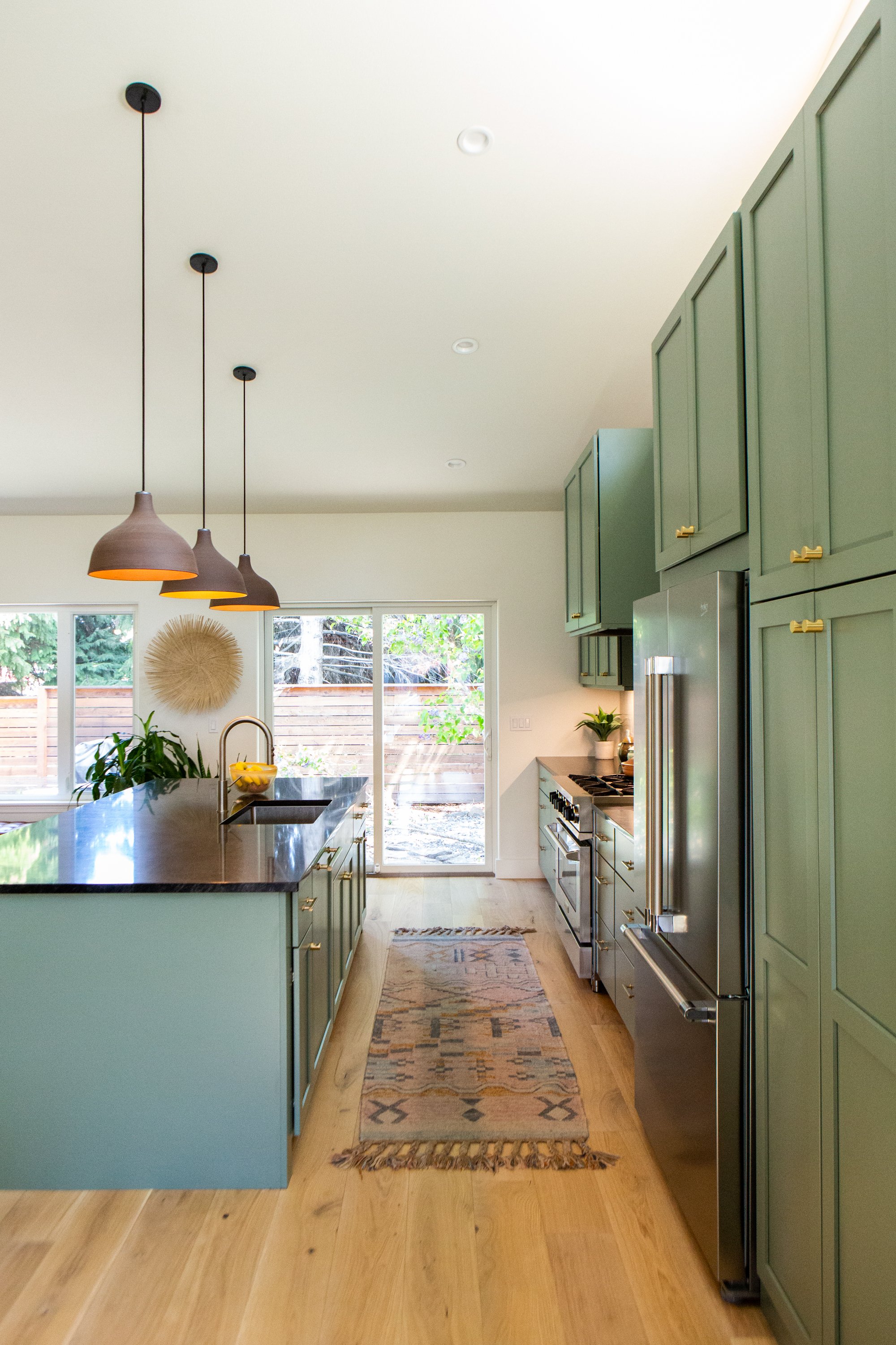 Kitchen with green cabinets, stainless steel refrigerator, light wood flooring, and a kitchen island with a sink. Three pendant lights hang from the ceiling over the island. Behind the island is a large window and sliding glass door showing an outdoo