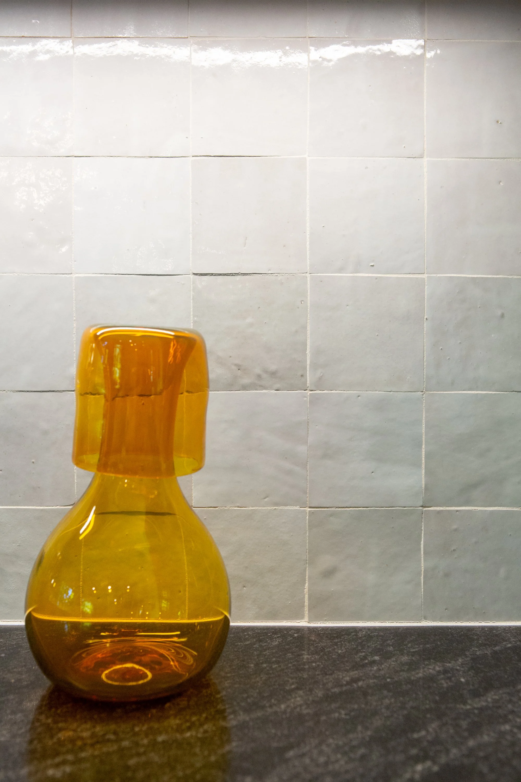 A yellow glass vase with a rounded base and narrow neck placed on a black surface in front of a gray tiled wall.